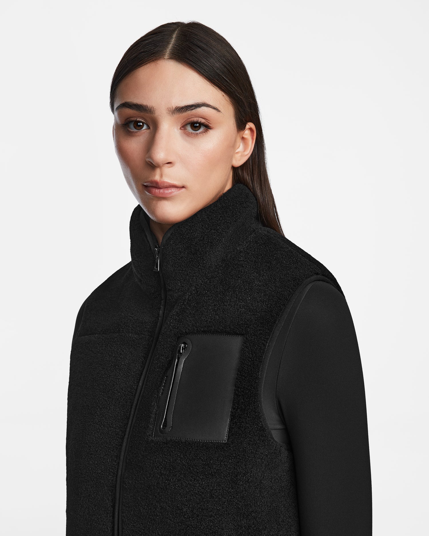 A woman with straight dark hair wears the Rudsak RUMMY - U524502 Black W unisex vest with a zippered chest pocket, standing against a plain white background and looking directly at the camera with a neutral expression.