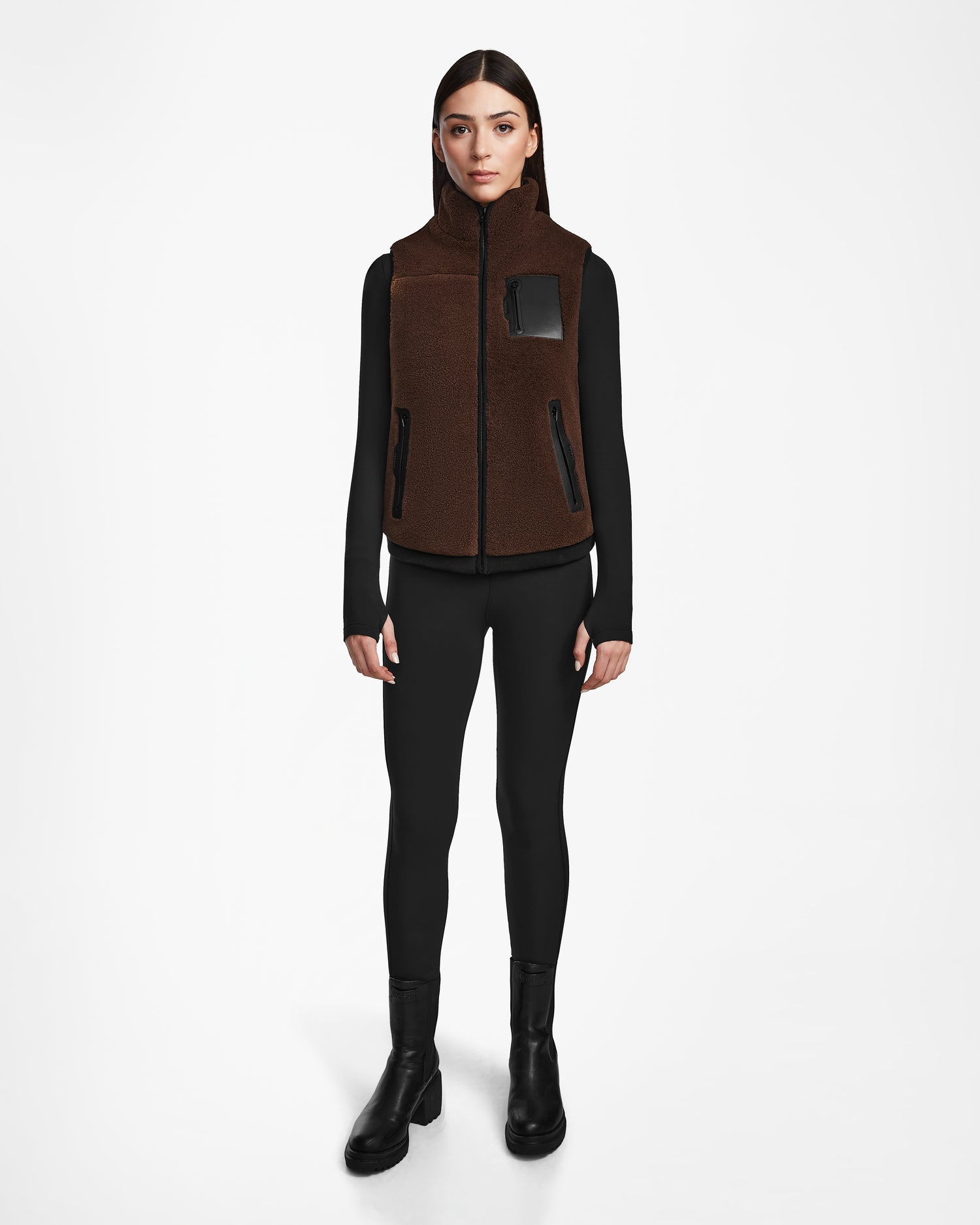 A woman faces forward wearing the Rudsak RUMMY - U524502 Espresso W unisex teddy zip-up vest with black details over a black long-sleeve top, leggings, and ankle boots, set against a plain white background.