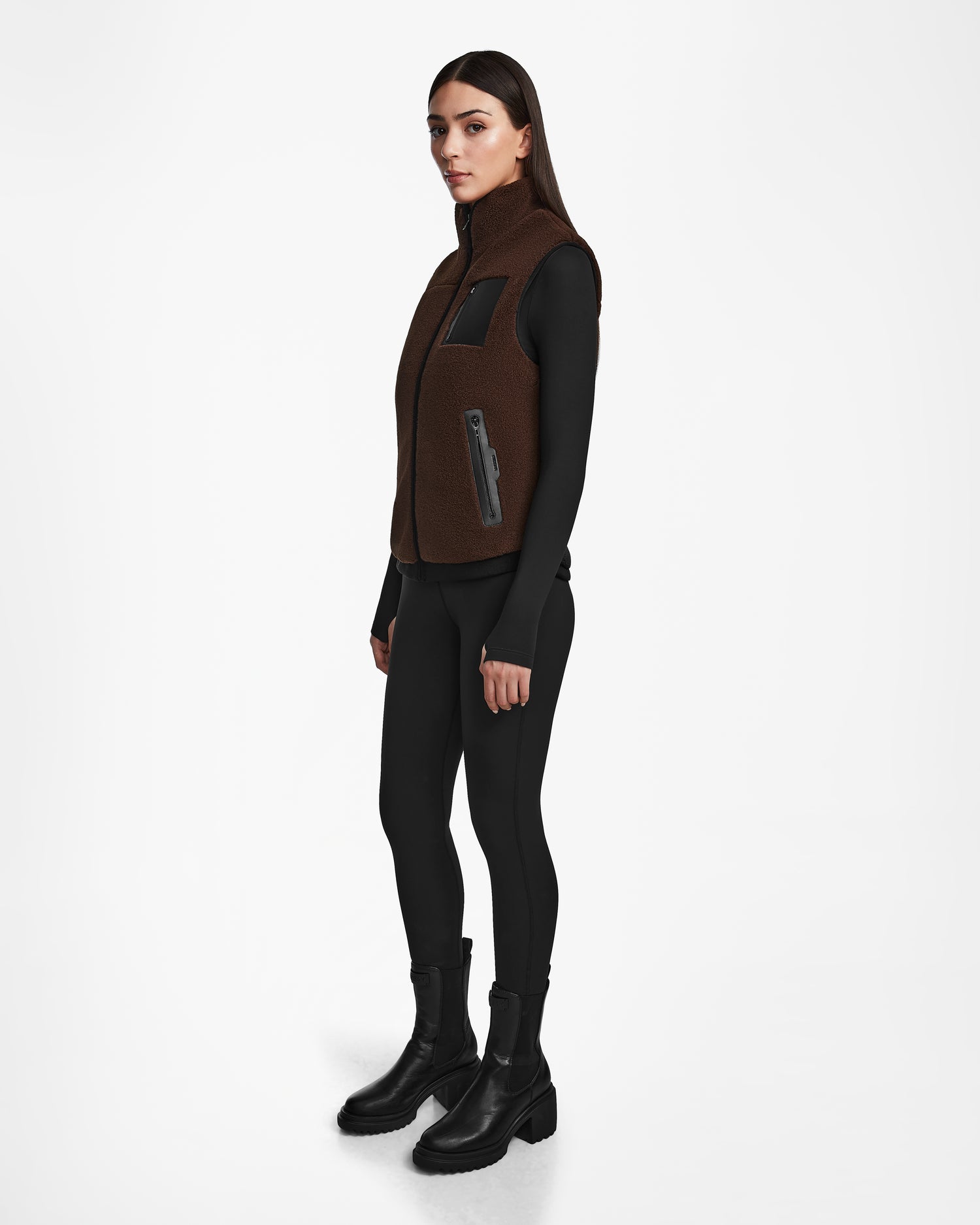 A woman wears the Rudsak RUMMY - U524502 Espresso W vest over a black long-sleeve top, black leggings, and ankle boots. She stands against a white background, facing slightly to the side with a neutral expression.