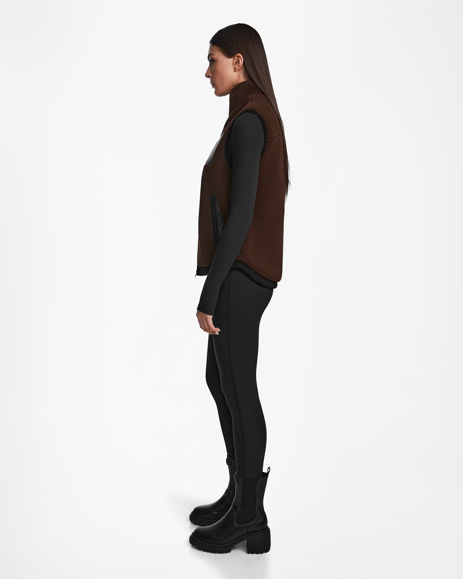 A woman in profile wears the Rudsak RUMMY - U524502 Espresso W unisex teddy vest over a black long-sleeve top, leggings, and ankle boots against a plain white background.