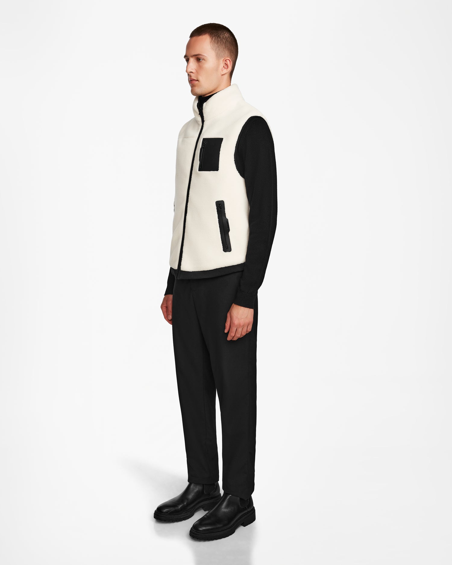 A man stands against a white background wearing the Rudsak RUMMY - U524502 Iceland M unisex polar fleece vest with black pockets and trim—an essential layering piece paired with a black long-sleeve shirt, pants, and shoes.
