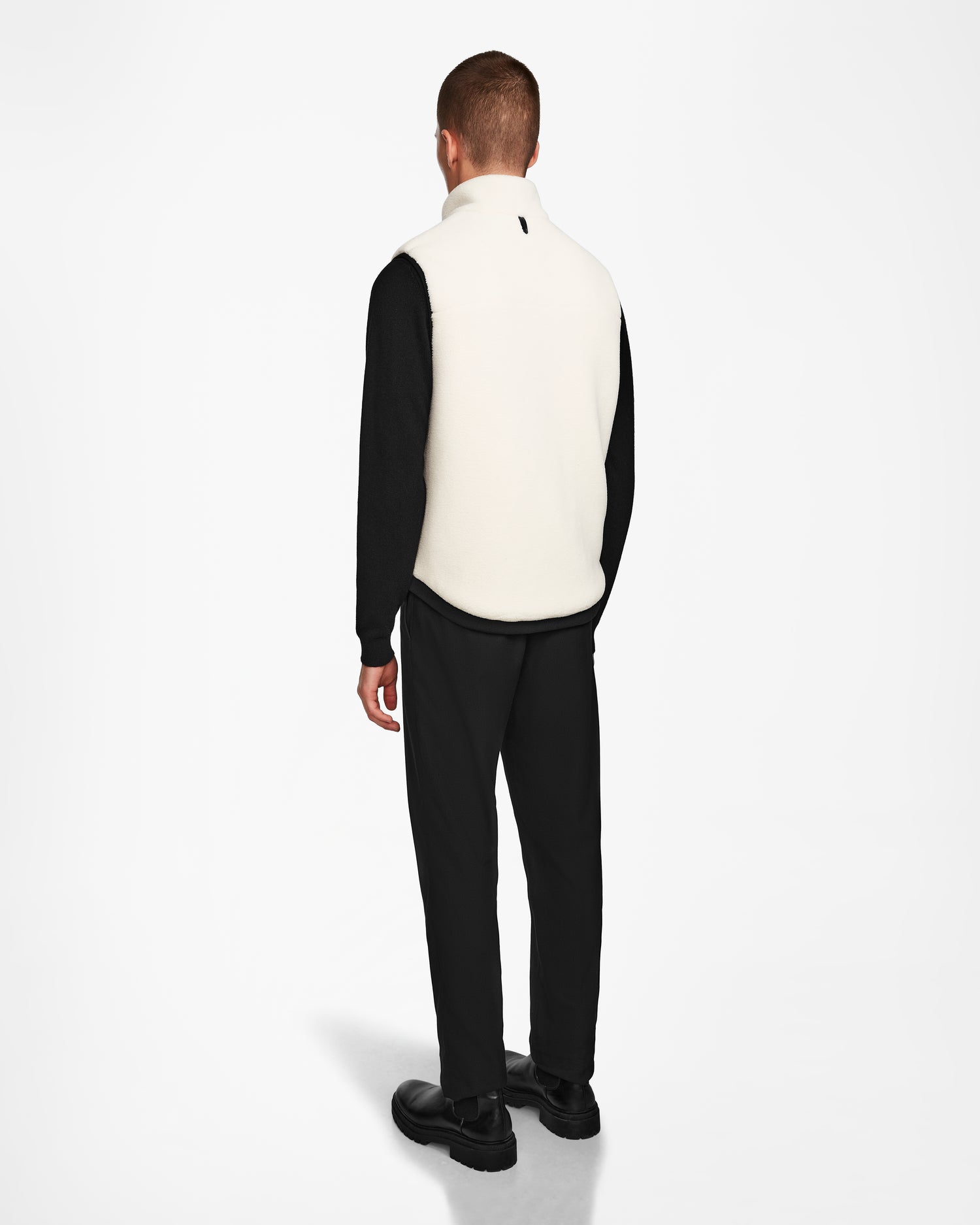A man faces away, wearing the Rudsak RUMMY - U524502 Iceland M cream unisex polar fleece vest over a black shirt and pants with black shoes, set against a plain white background—an ideal layering essential.