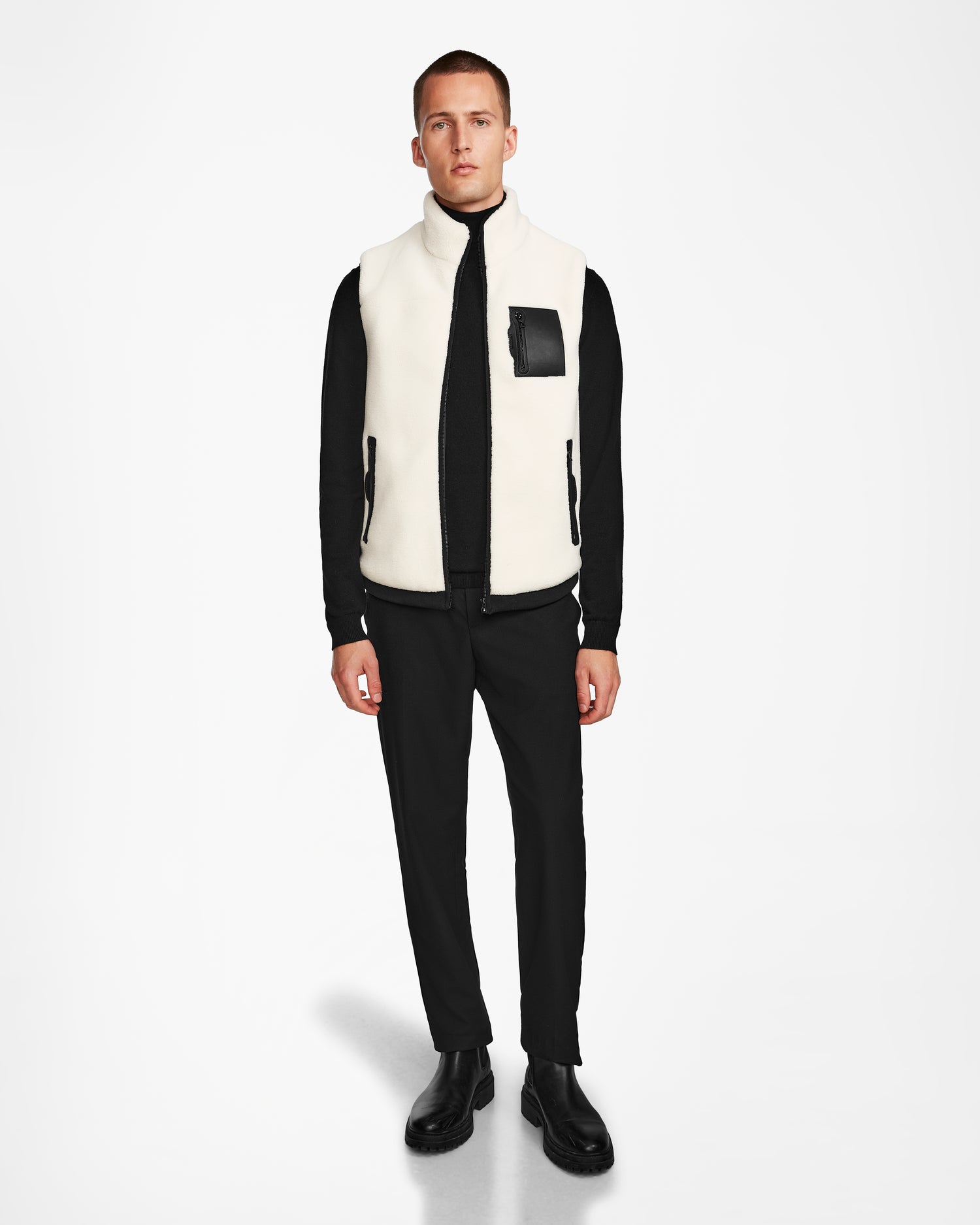 A man stands against a white background wearing the Rudsak RUMMY - U524502 Iceland M unisex polar fleece vest with a black pocket, paired with a black zip-up top, pants, and shoes—showcasing a versatile layering piece for all seasons.