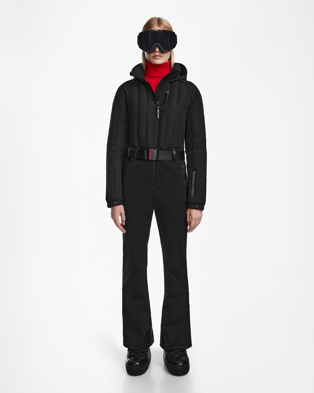 A person faces forward wearing the Rudsak SAGE - 8124782 Black womens ski suit with RECCO rescue tech, large black ski goggles, black boots, and a red turtleneck, set against a plain white background.