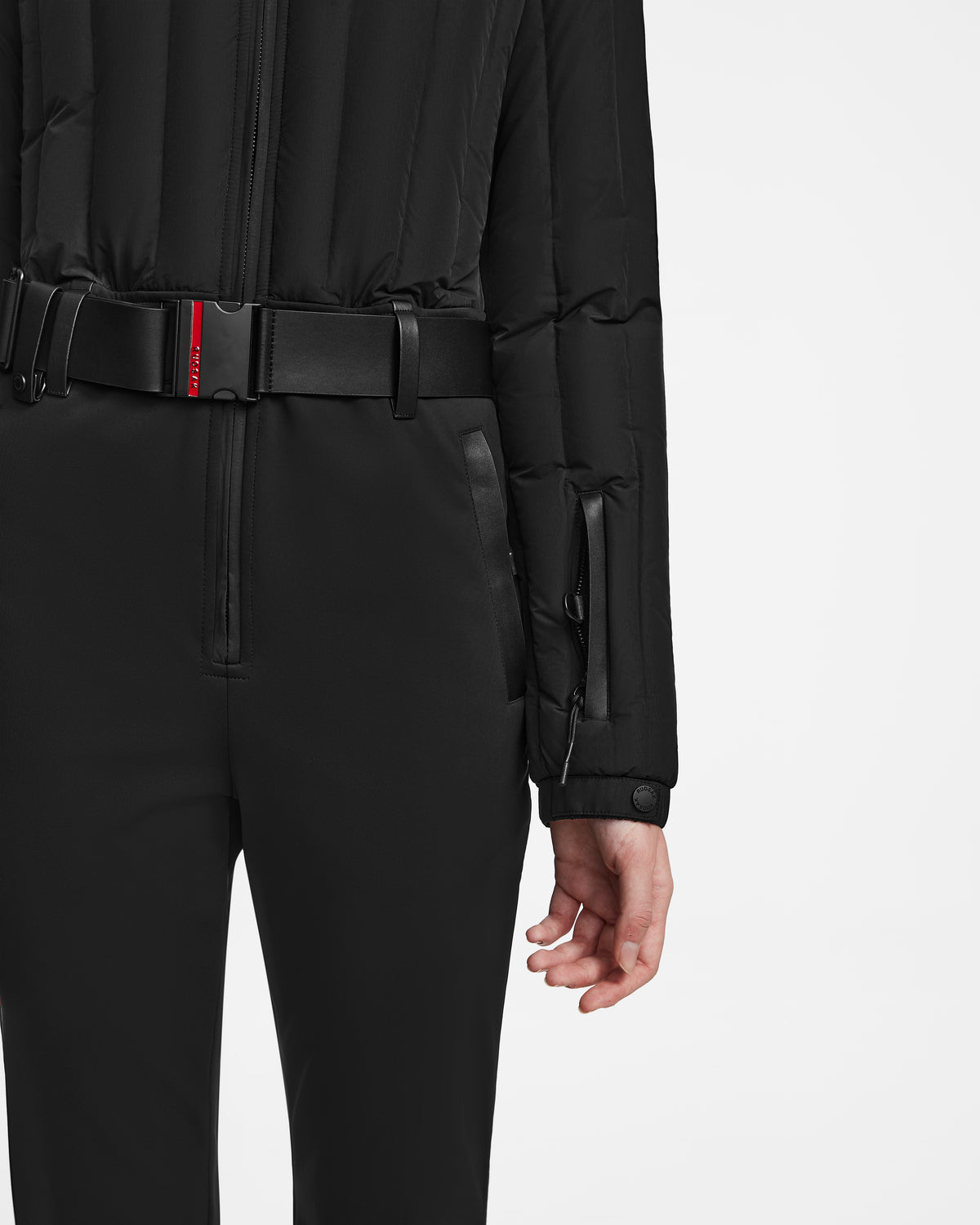 A person is photographed from the back wearing the Rudsak SAGE - 8124782 Black womens ski suit, featuring high-waisted pants, a quilted jacket, and a black belt with a small red detail, right hand resting by their side.