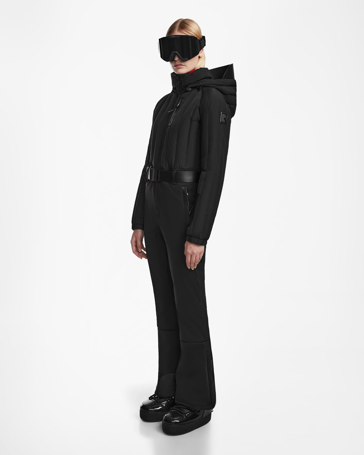 A woman faces forward in the Rudsak SAGE - 8124782 Black women’s ski suit with a hood, wide black belt, gloves, large ski goggles, and shiny black boots. Her blonde hair is tied back. The suit features advanced RECCO rescue technology. White background.