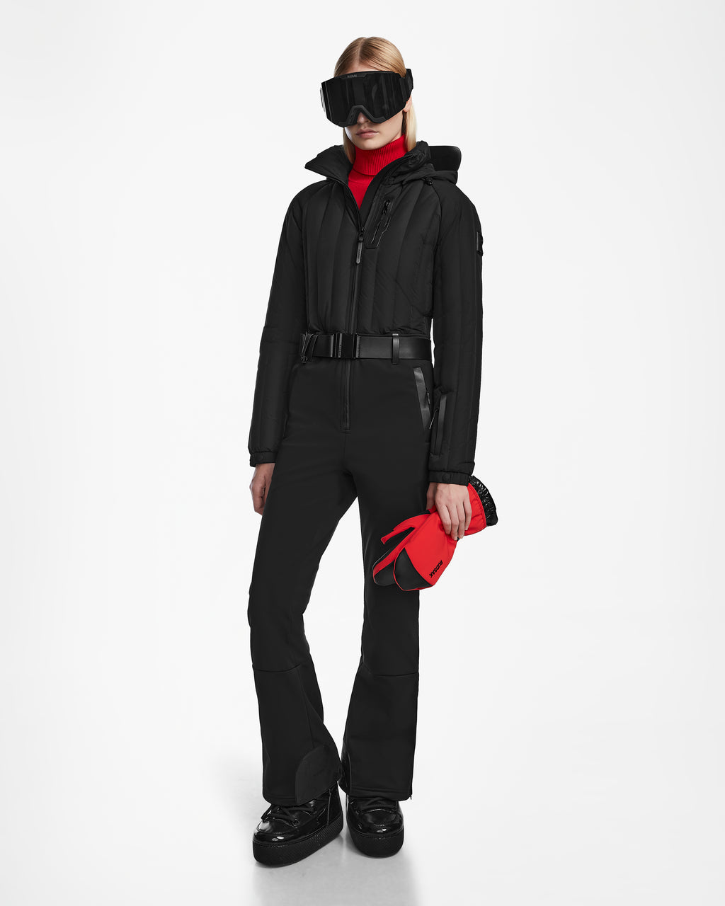 A person wearing the Rudsak SAGE - 8124782 Black one-piece ski suit stands against a plain white background, paired with black snow boots, black ski goggles, a red turtleneck, and holding red ski gloves.