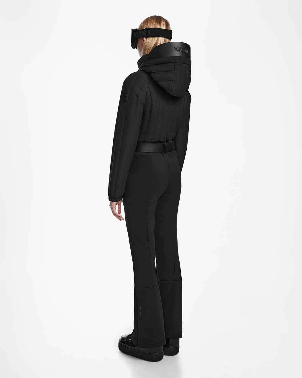 A person with long blonde hair, wearing the Rudsak SAGE - 8124782 Black women’s ski suit featuring RECCO rescue technology and a black ski visor, stands facing away against a plain white background.