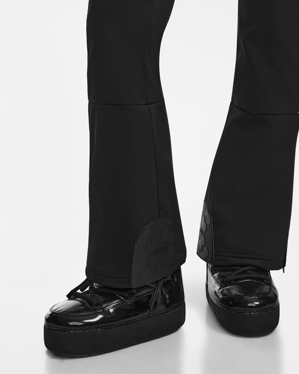 Close-up of Rudsaks SAGE - 8124782 Black zippered flared pants over glossy black platform shoes, inspired by womens ski suits. The look embodies modern winter style with sporty sophistication on a white background.