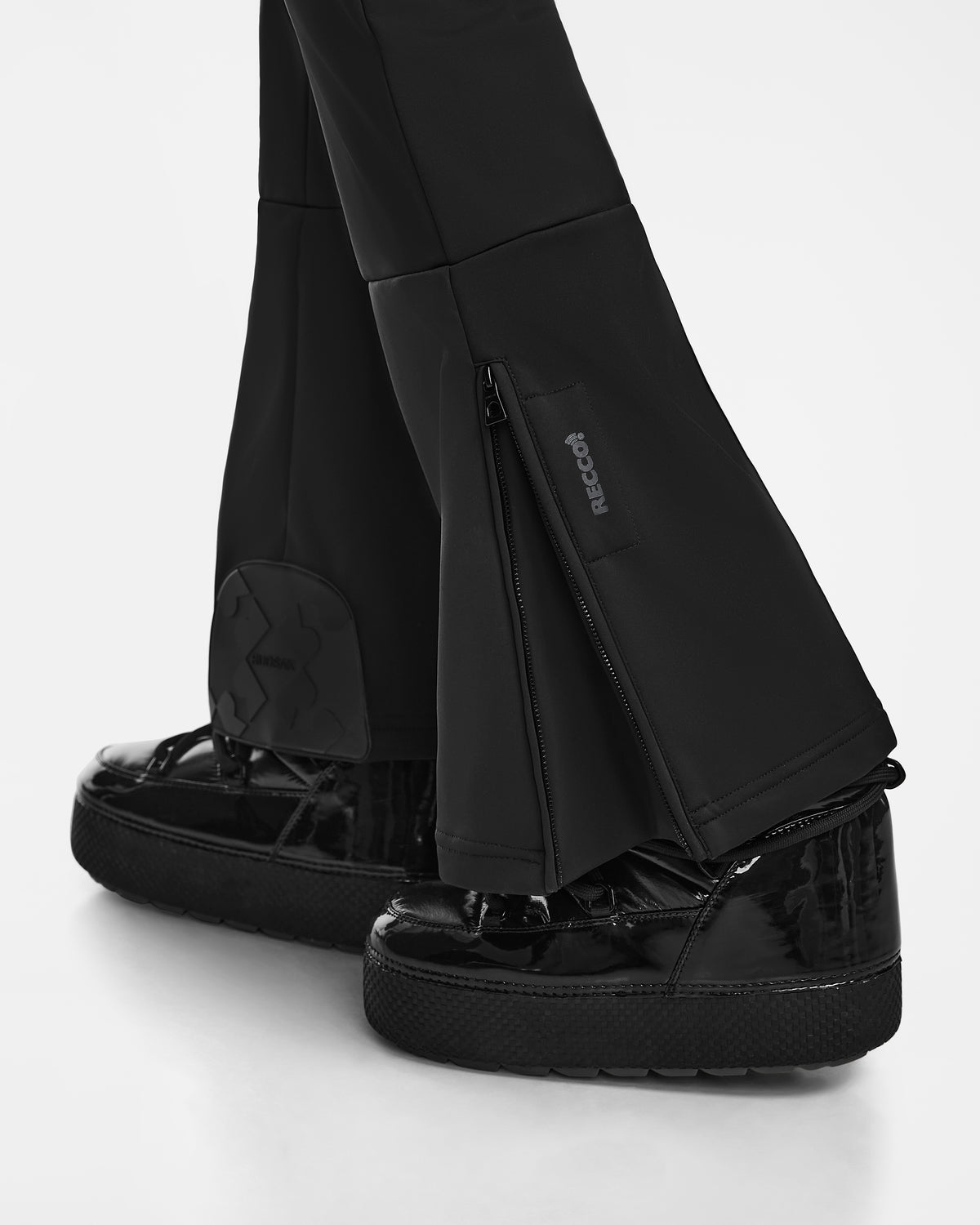 Close-up of a person wearing Rudsak SAGE - 8124782 Black pants with zippers and the RECCO logo, highlighting integrated rescue technology. The black pants partially cover platform shoes against a plain white background.