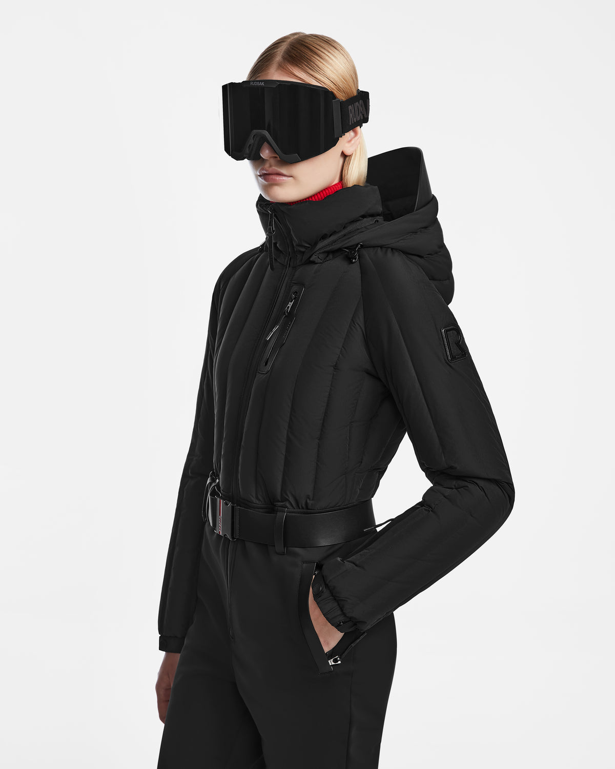 A person wearing the Rudsak SAGE - 8124782 Black technical performance ski suit, featuring a hooded jacket, wide belt, and large black ski goggles, stands against a plain white background.