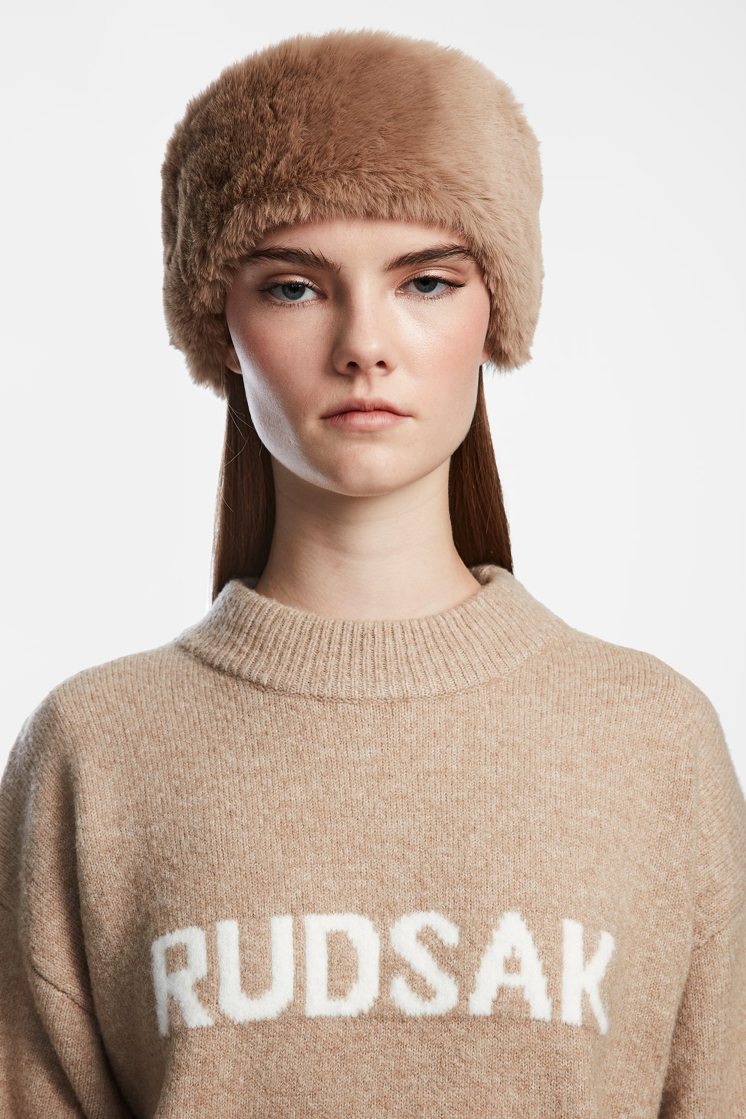 A woman with straight brown hair wears the SANDIE - 8423507 Camel faux fur hat and a beige Rudsak sweater, standing against a white background. Her stylish winter accessory by Rudsak adds cozy elegance to her look.