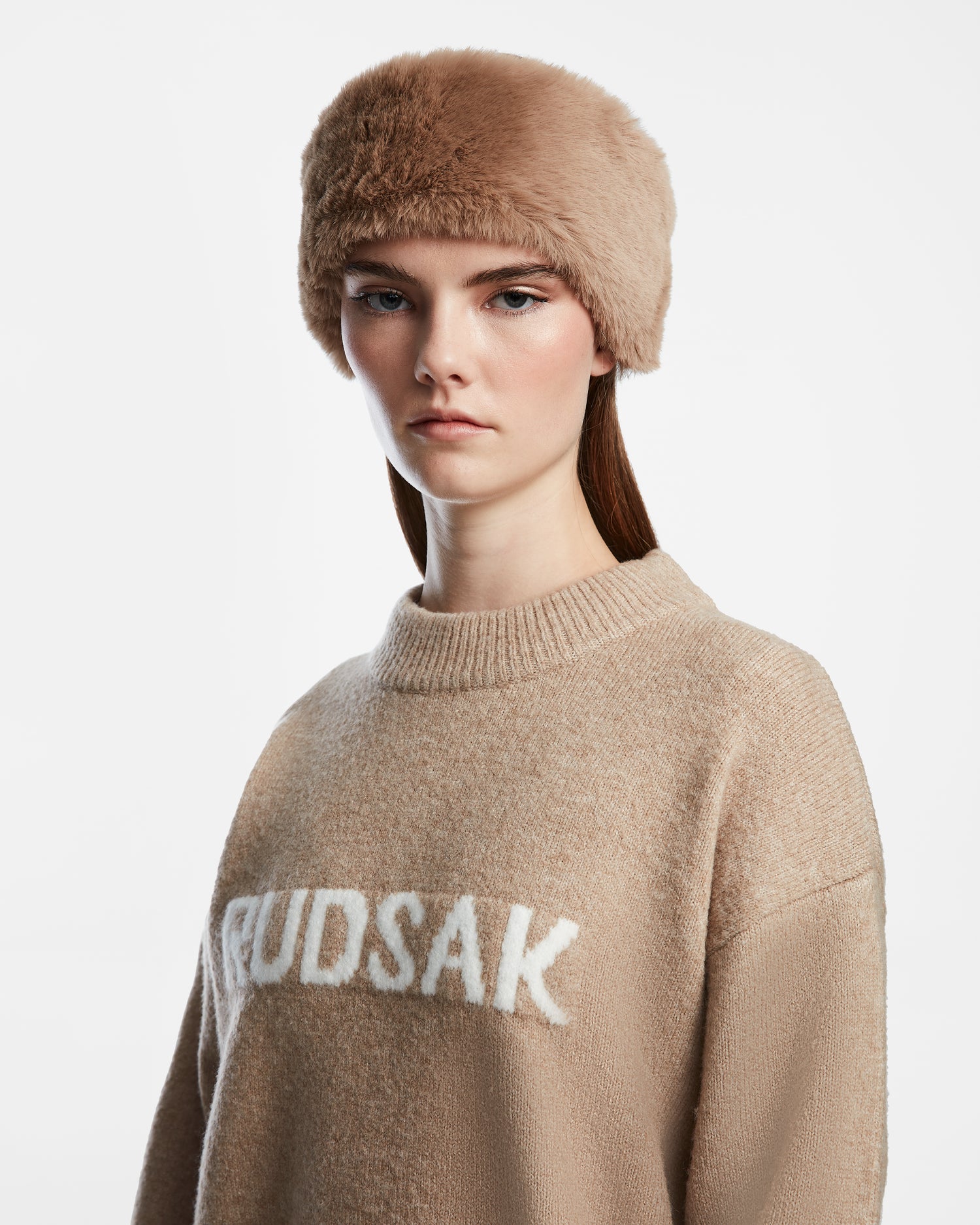 A woman stands against a white background, wearing the Rudsak SANDIE - 8423507 Camel faux fur headband and a beige sweater with RUDSAK on it, highlighting this chic winter accessory.