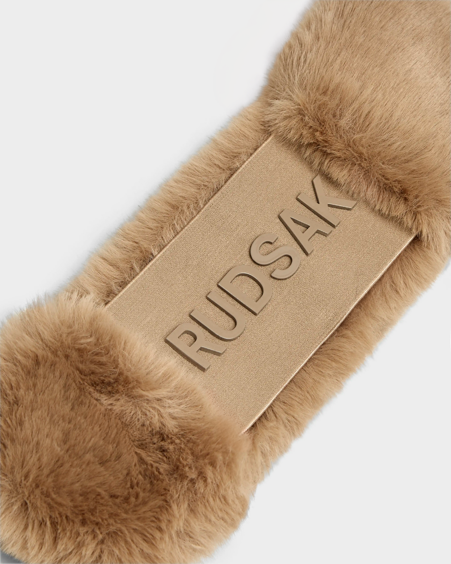 A close-up of the SANDIE - 8423507 Camel, a tan faux fur headband by Rudsak, featuring the brand embossed on a smooth rectangular panel at the center—a chic winter accessory for women.