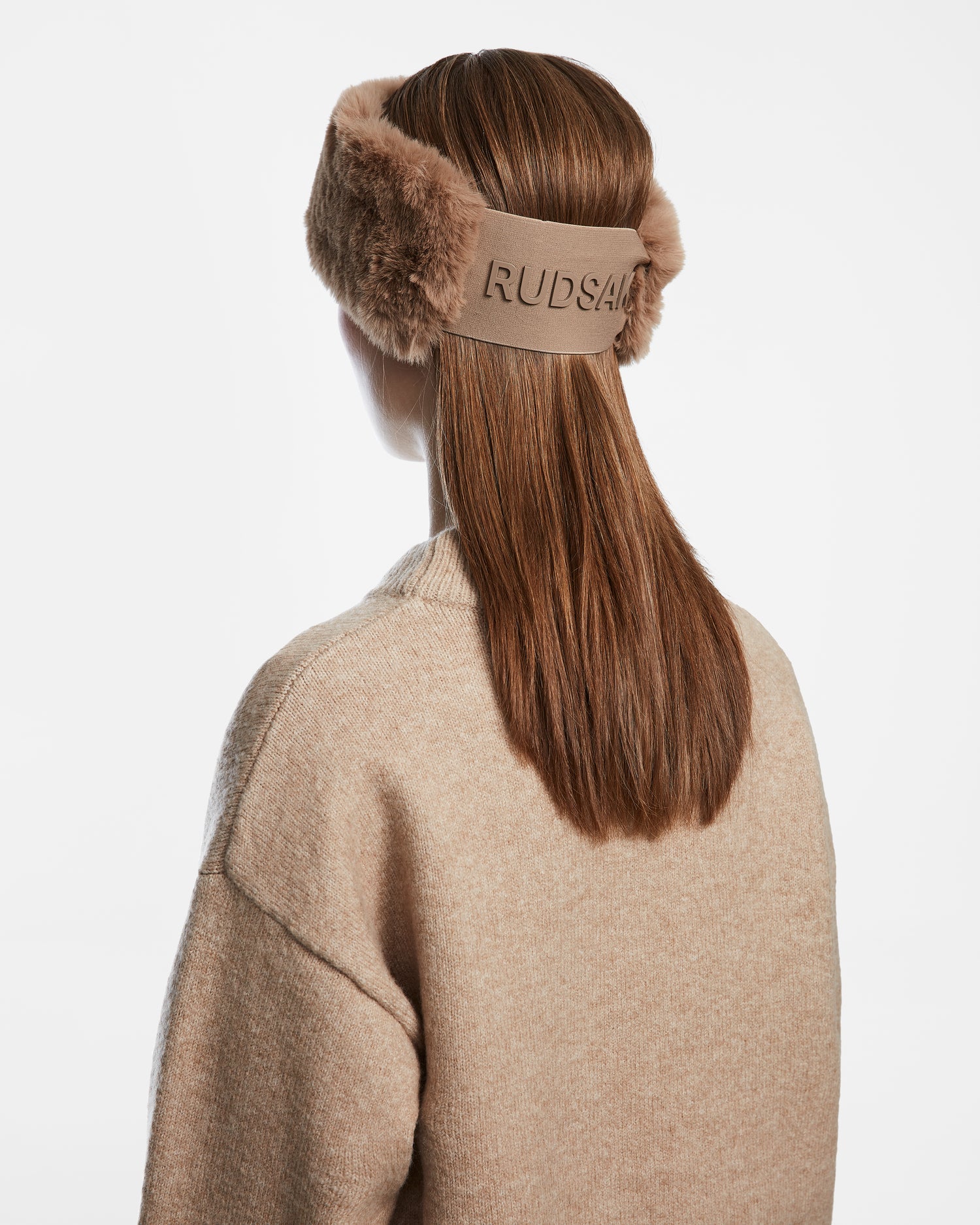 A woman with straight brown hair wears a tan sweater and the Rudsak SANDIE - 8423507 Camel faux fur headband, embroidered with the brand name—a chic winter accessory—against a white background.