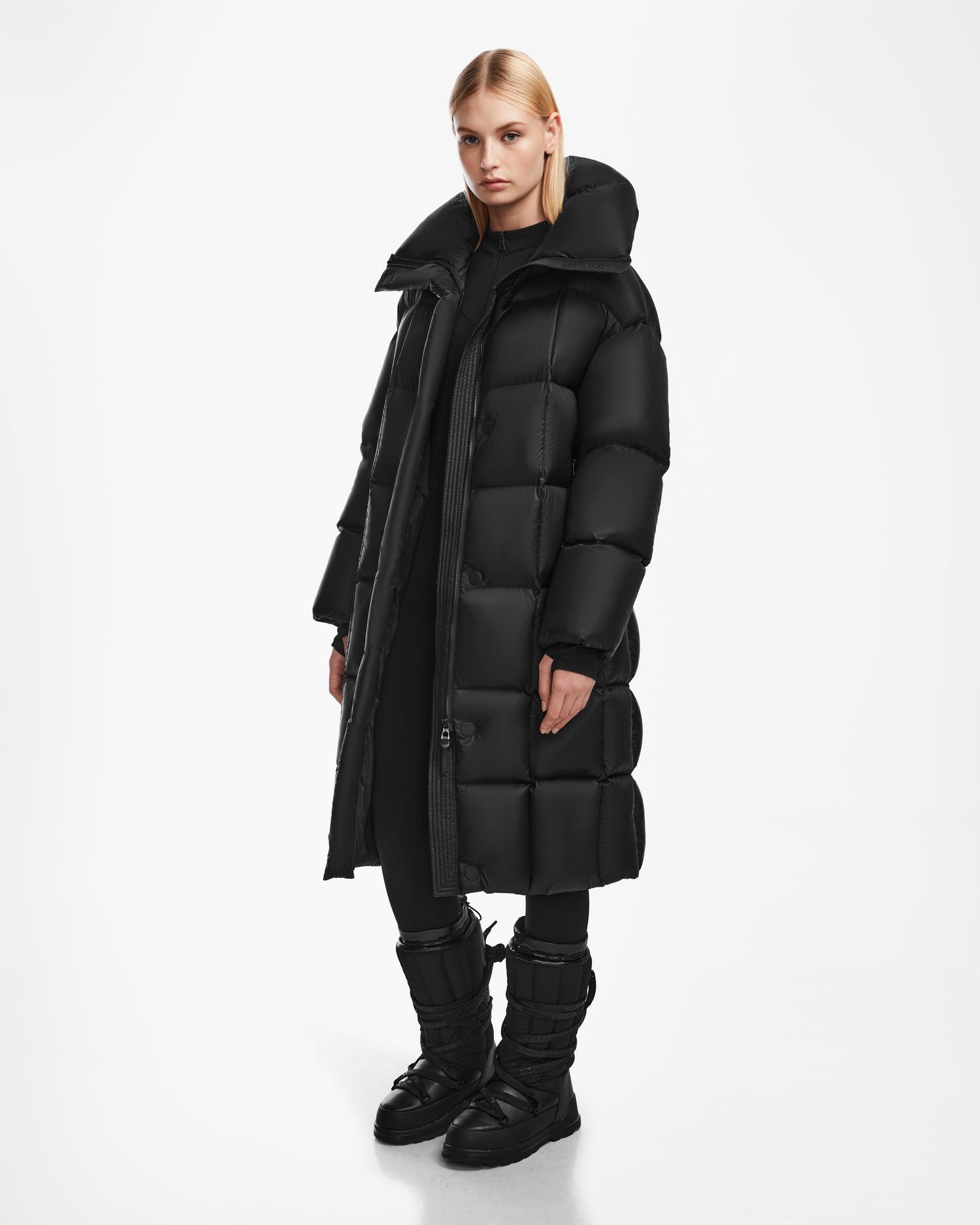 A person with straight blonde hair wears the Rudsak SASH - 8124536 Black womens long down puffer coat, paired with black pants and black boots, standing against a plain white background.