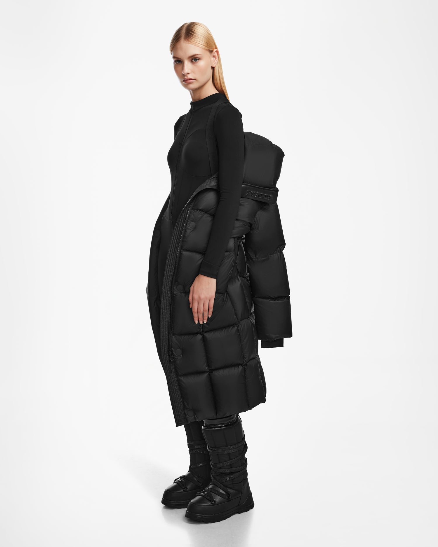 A woman with straight blonde hair stands in profile, wearing a fitted black outfit, black boots, and the Rudsak SASH - 8124536 Black womens long down puffer coat draped over one shoulder against a plain white background.