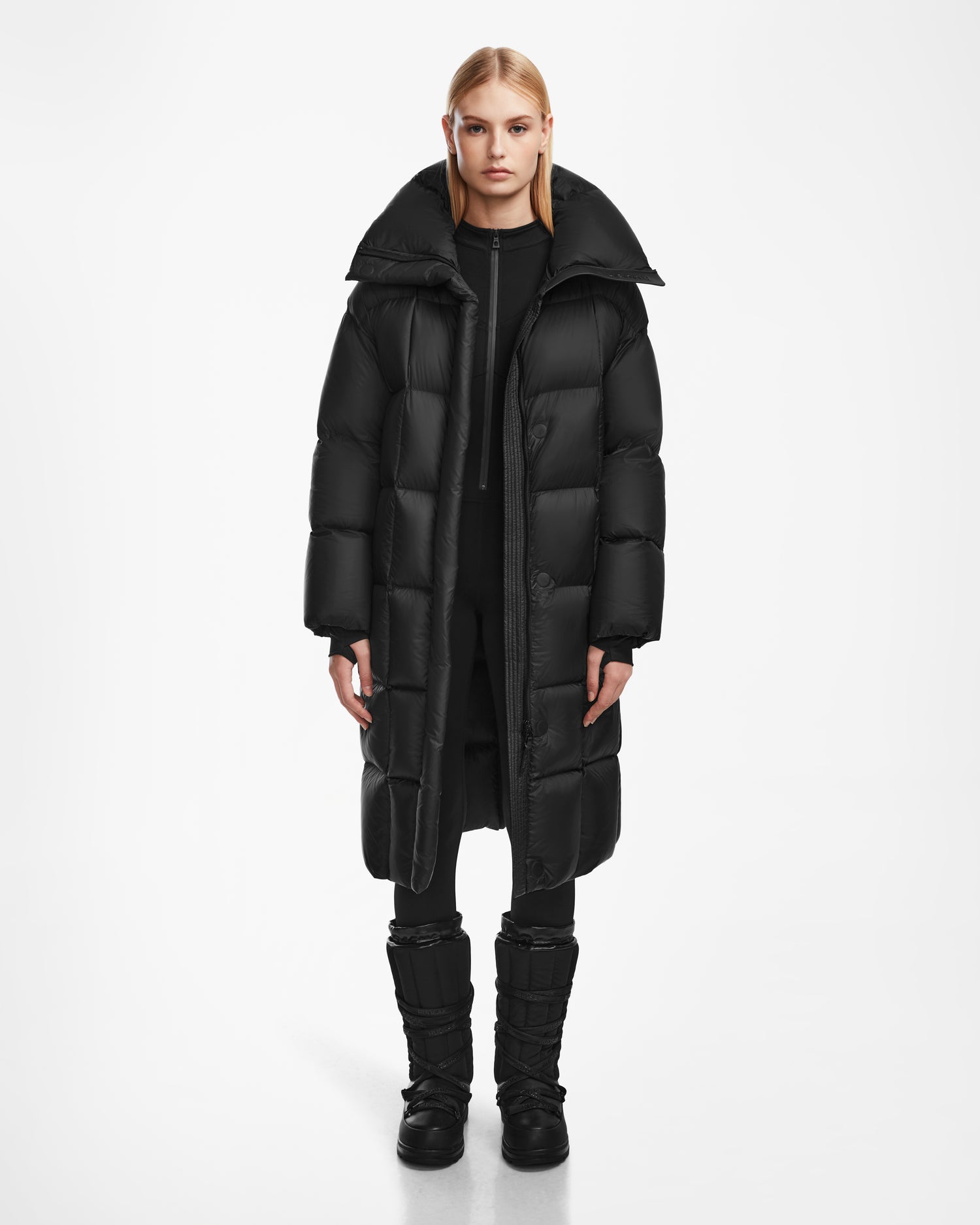 A person faces forward against a plain white background, wearing the Rudsak SASH - 8124536 Black womens long down puffer coat, paired with black pants and black boots.