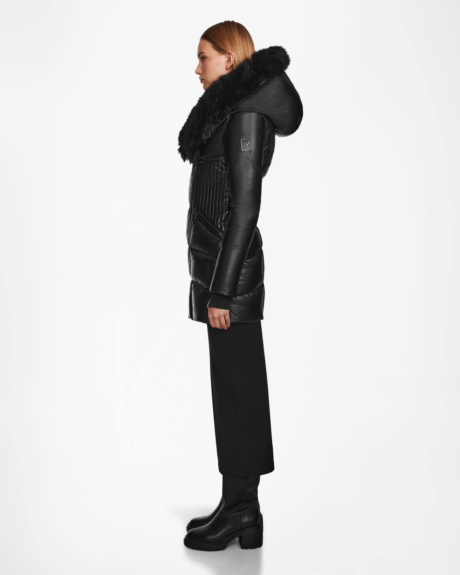 A woman in profile wears the Rudsak SHAUNA X - 8122535 BLACK women’s puffer coat with RDS certified down and a large fur-trimmed hood, paired with wide-leg black pants and heeled boots, set against a plain white background.