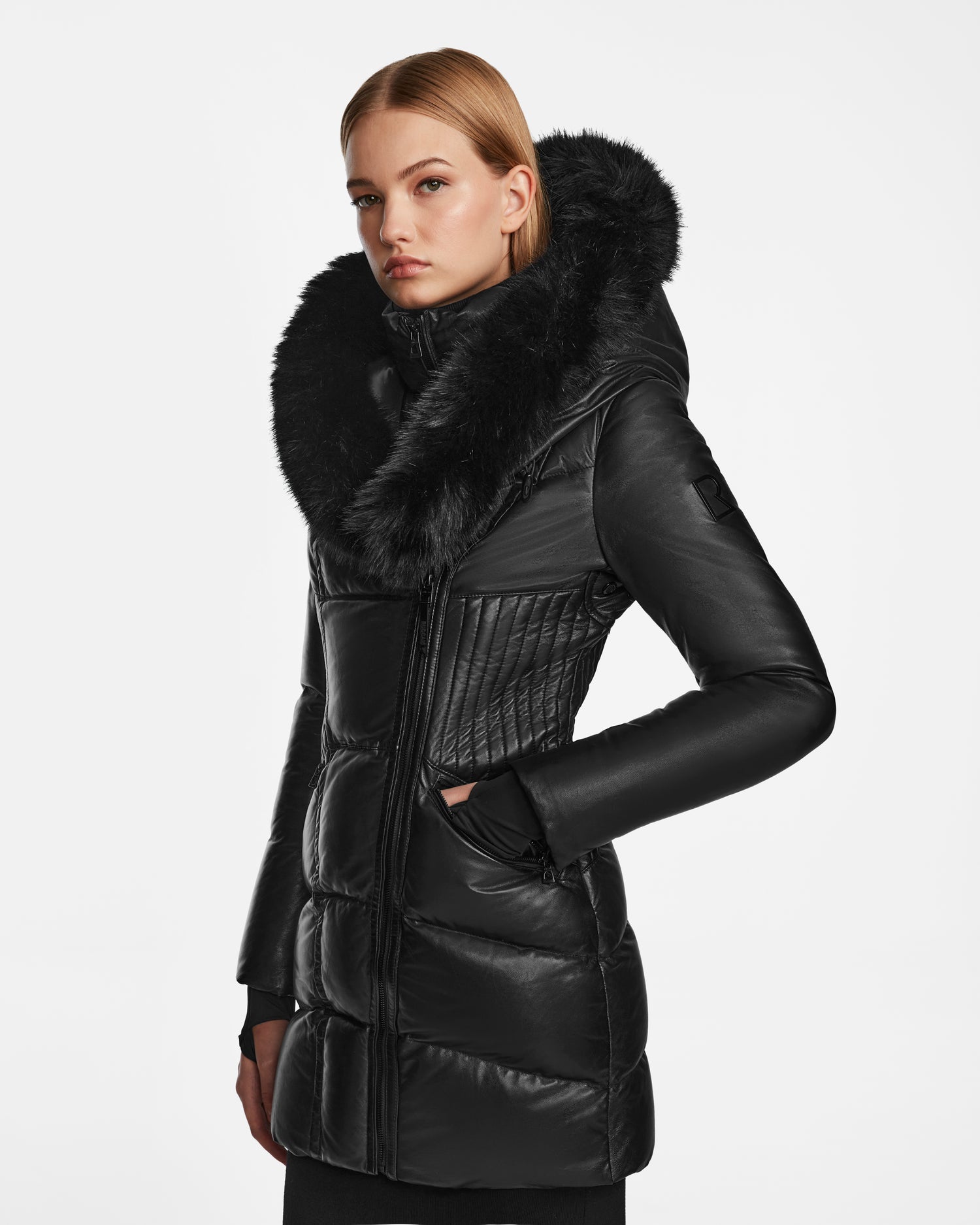 A woman stands with one hand in her pocket, wearing the Rudsak SHAUNA X - 8122535 BLACK quilted women’s puffer hooded winter coat with fur trim, looking slightly to the side against a plain white background.