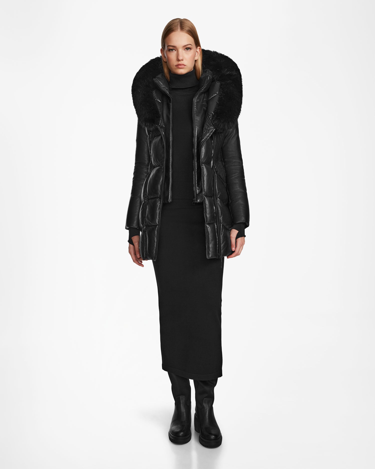 A woman wears the SHAUNA X - 8122535 BLACK puffer coat by Rudsak, featuring a large fur-trimmed hood, over a black turtleneck dress and boots, standing against a plain white background.