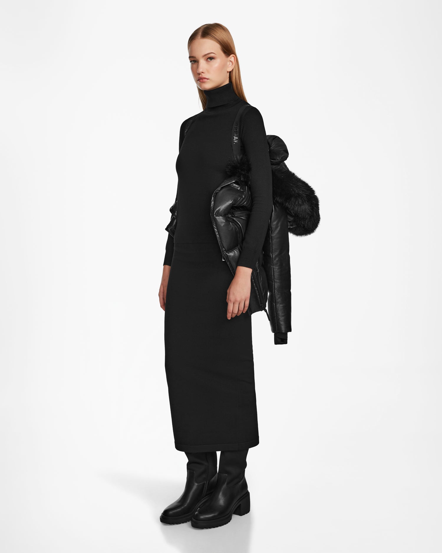 A woman stands against a white background wearing a black turtleneck, long skirt, black boots, and the Rudsak SHAUNA X - 8122535 BLACK RDS certified down women’s puffer jacket with fur trim draped over her shoulders.