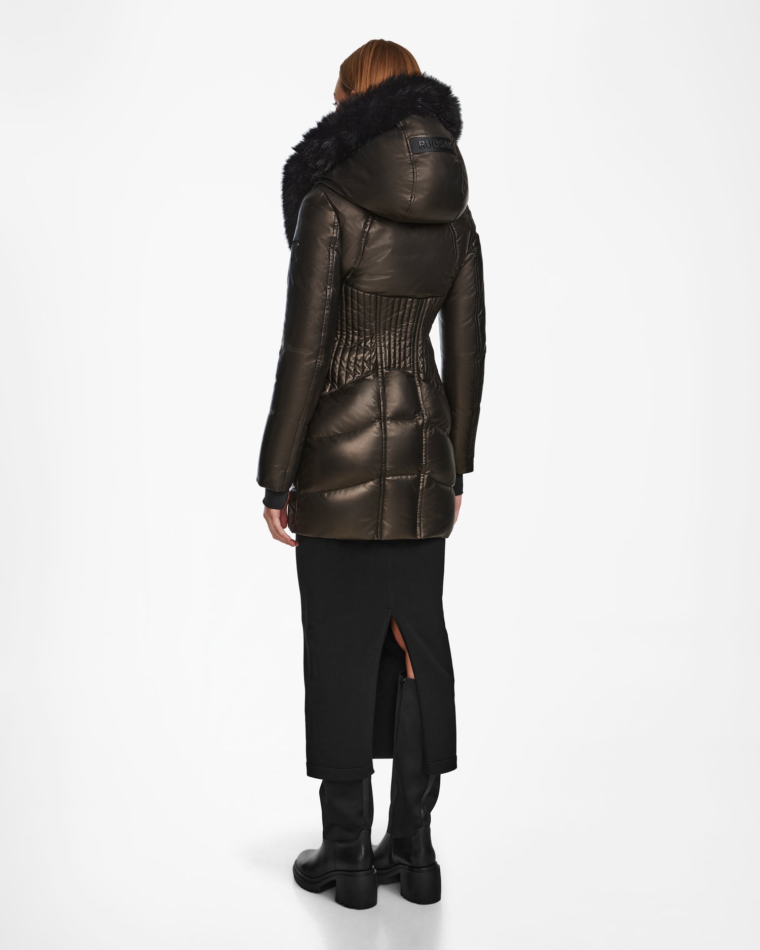 A person faces away in the Rudsak SHAUNA X - 8122535 BLACK women’s puffer coat with RDS certified down and a furry hood, styled with a black skirt featuring a back slit and black boots, set against a plain white background.