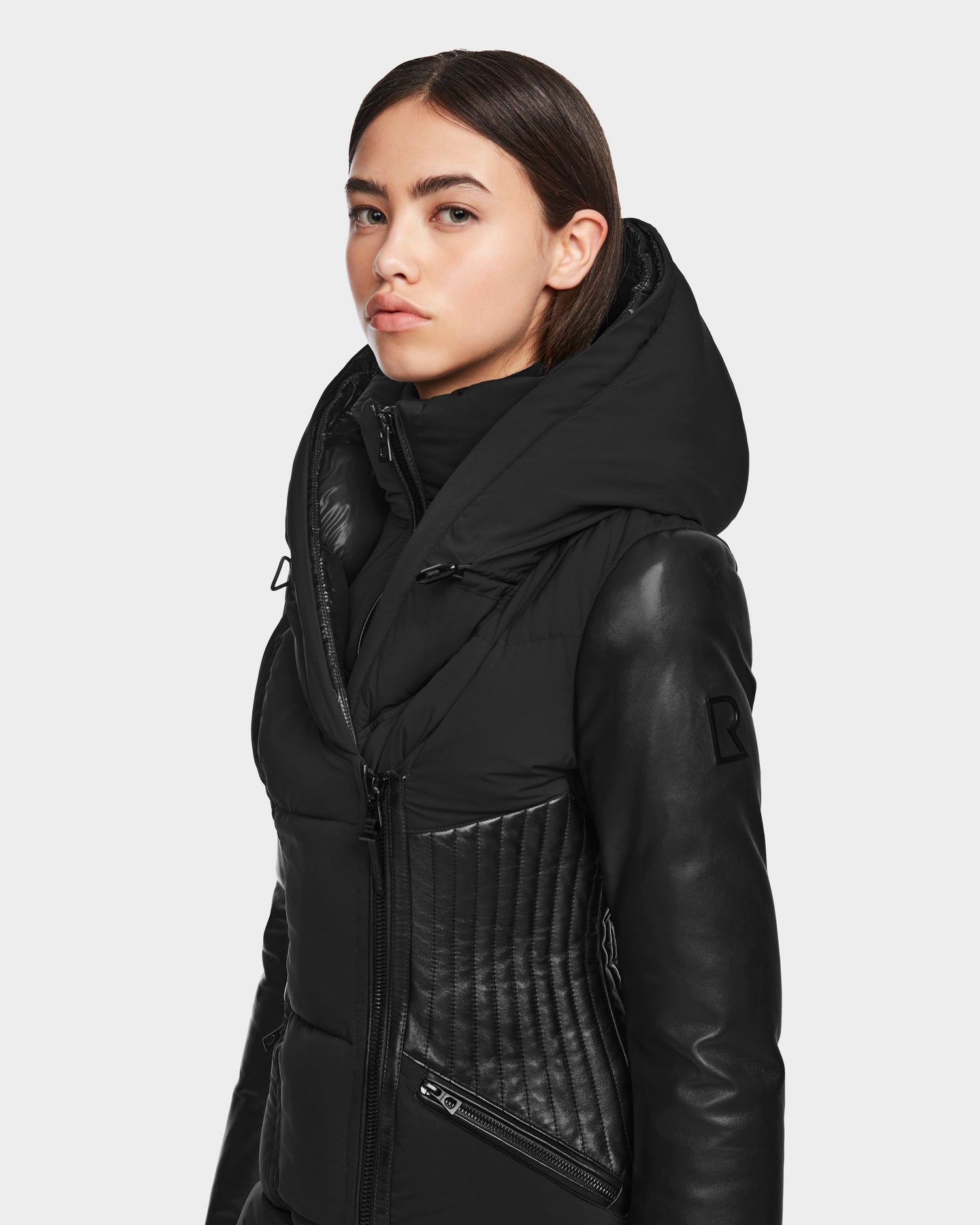 A woman with straight dark hair wears the RUDSAK SHAUNA - 8122586 BLACK quilted down puffer jacket with leather sleeves and a large hood, standing against a light background—a stylish, RDS-certified winter coat.