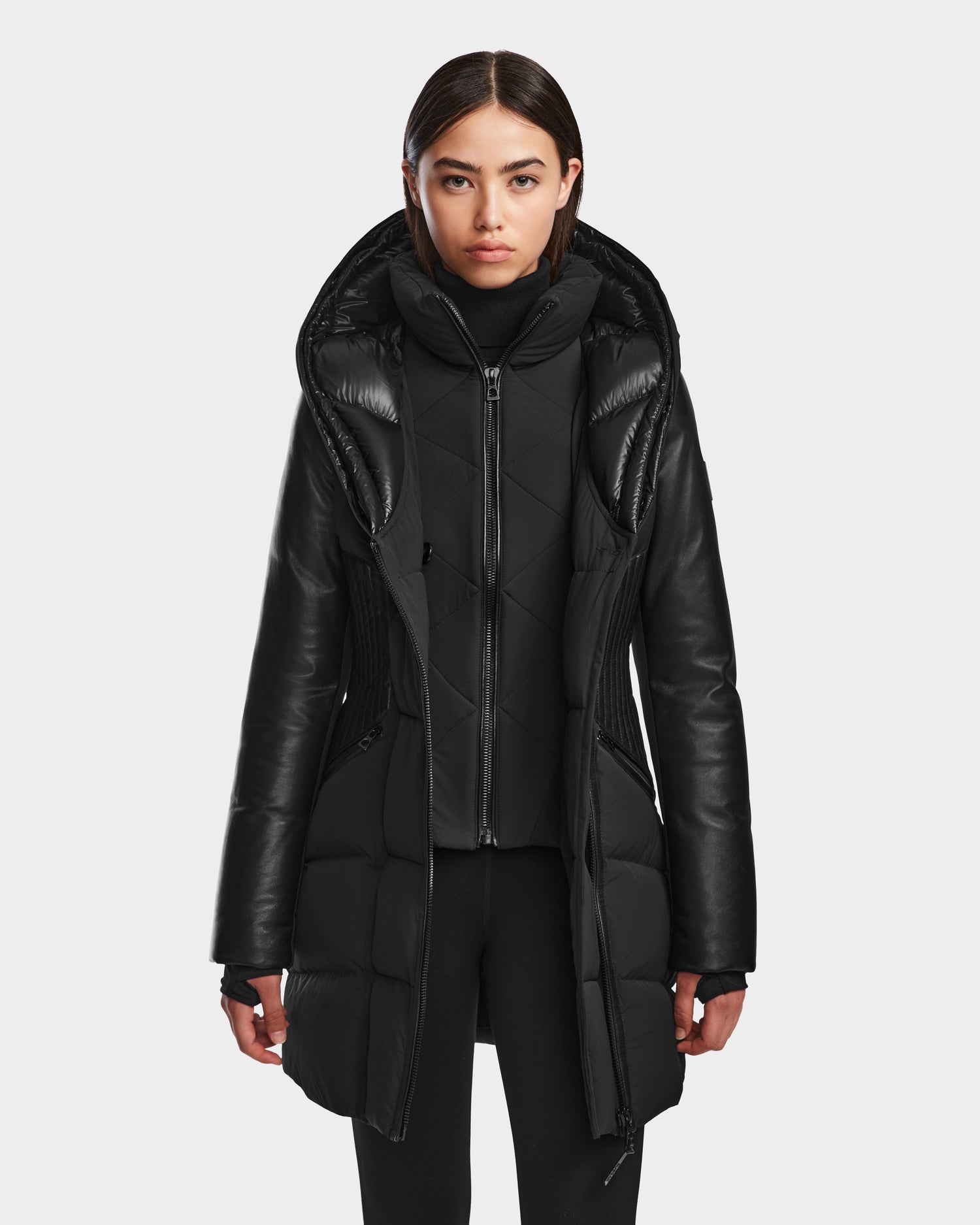 A woman with straight brown hair wears the RUDSAK SHAUNA - 8122586 BLACK hooded quilted down puffer coat, zipped up over black pants against a light gray background, its shiny and matte textures perfect for cold weather.