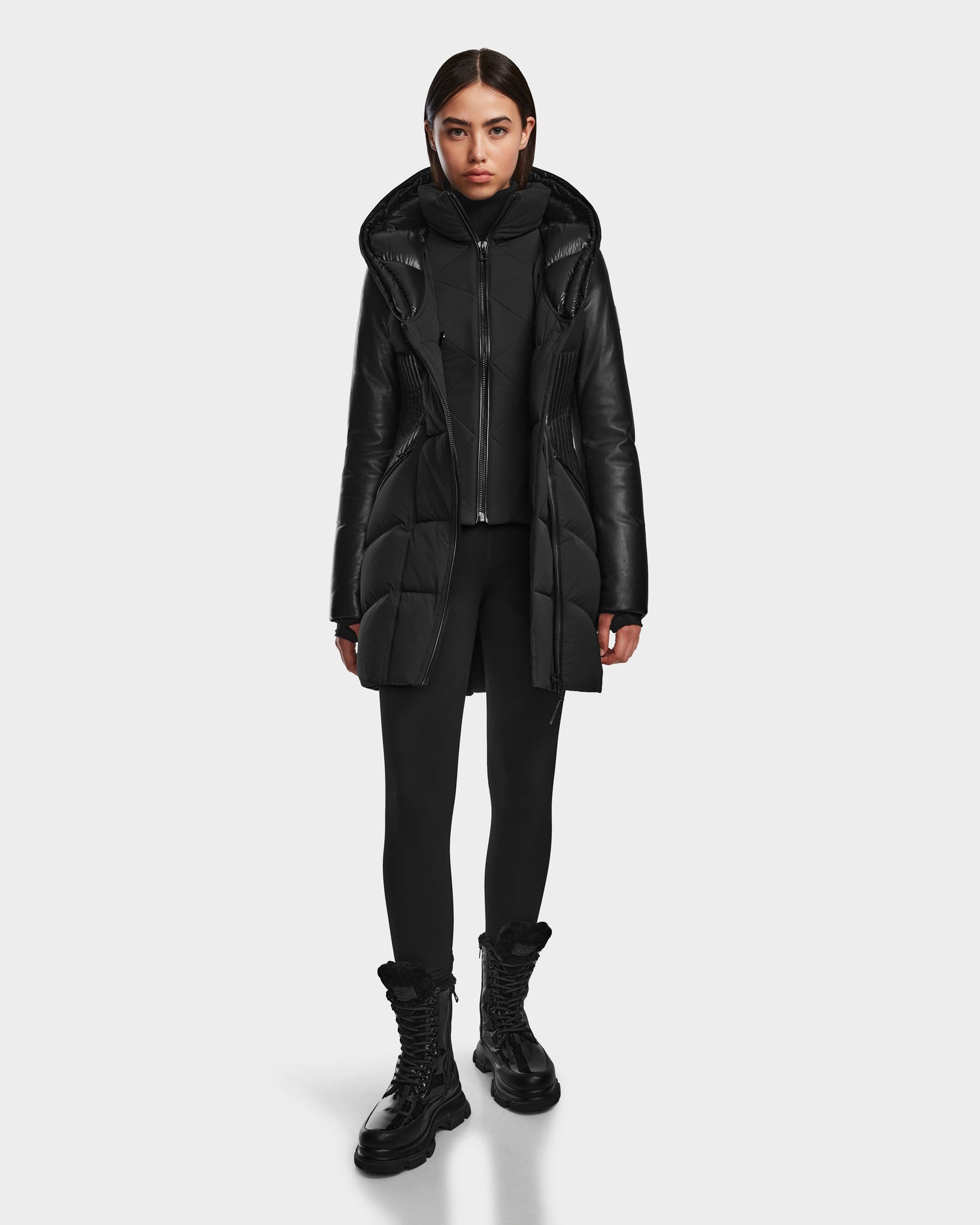 A woman faces forward in the SHAUNA - 8122586 BLACK winter coat by RUDSAK, paired with black leggings, a black top, and black lace-up boots against a plain white background.