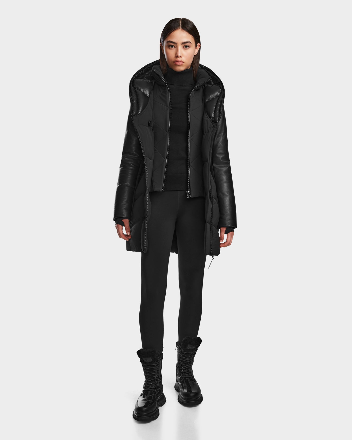 A woman faces forward in the SHAUNA - 8122586 BLACK quilted down puffer coat by RUDSAK, styled with a hood, black turtleneck, leggings, and lace-up boots—an effortless winter essential against a plain light background.