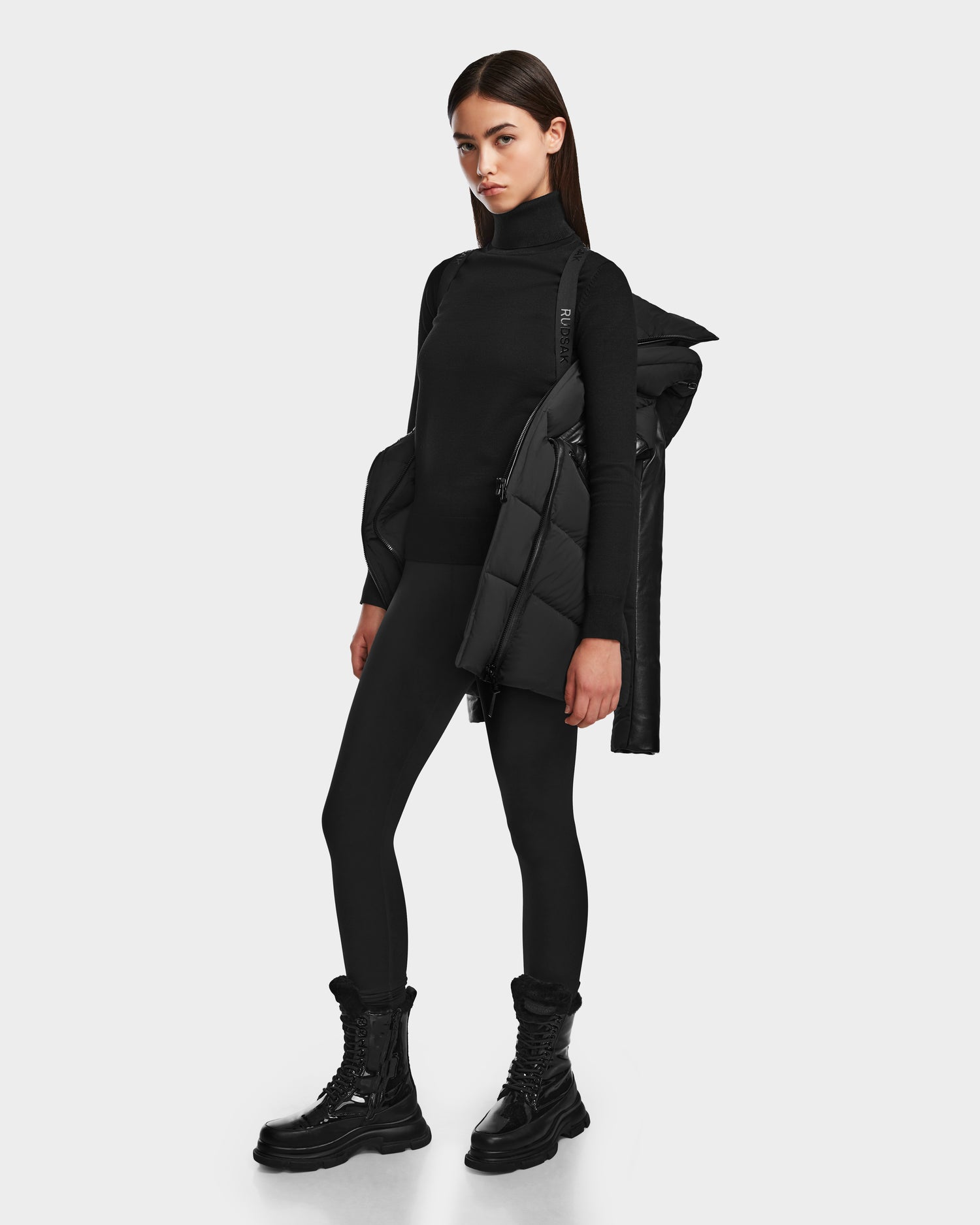 A woman poses confidently in the RUDSAK SHAUNA - 8122586 BLACK quilted down puffer jacket, paired with black leggings, a turtleneck, and boots—an essential winter look.