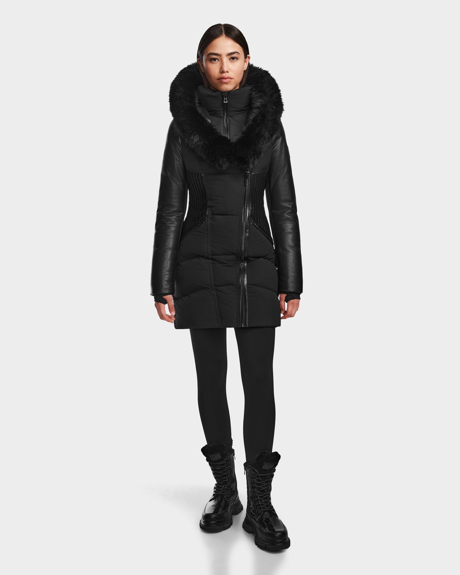 A woman faces forward wearing the RUDSAK SHAUNA - 8122586 BLACK long winter coat with a large fur-lined hood, paired with black leggings and lace-up boots against a plain white background.