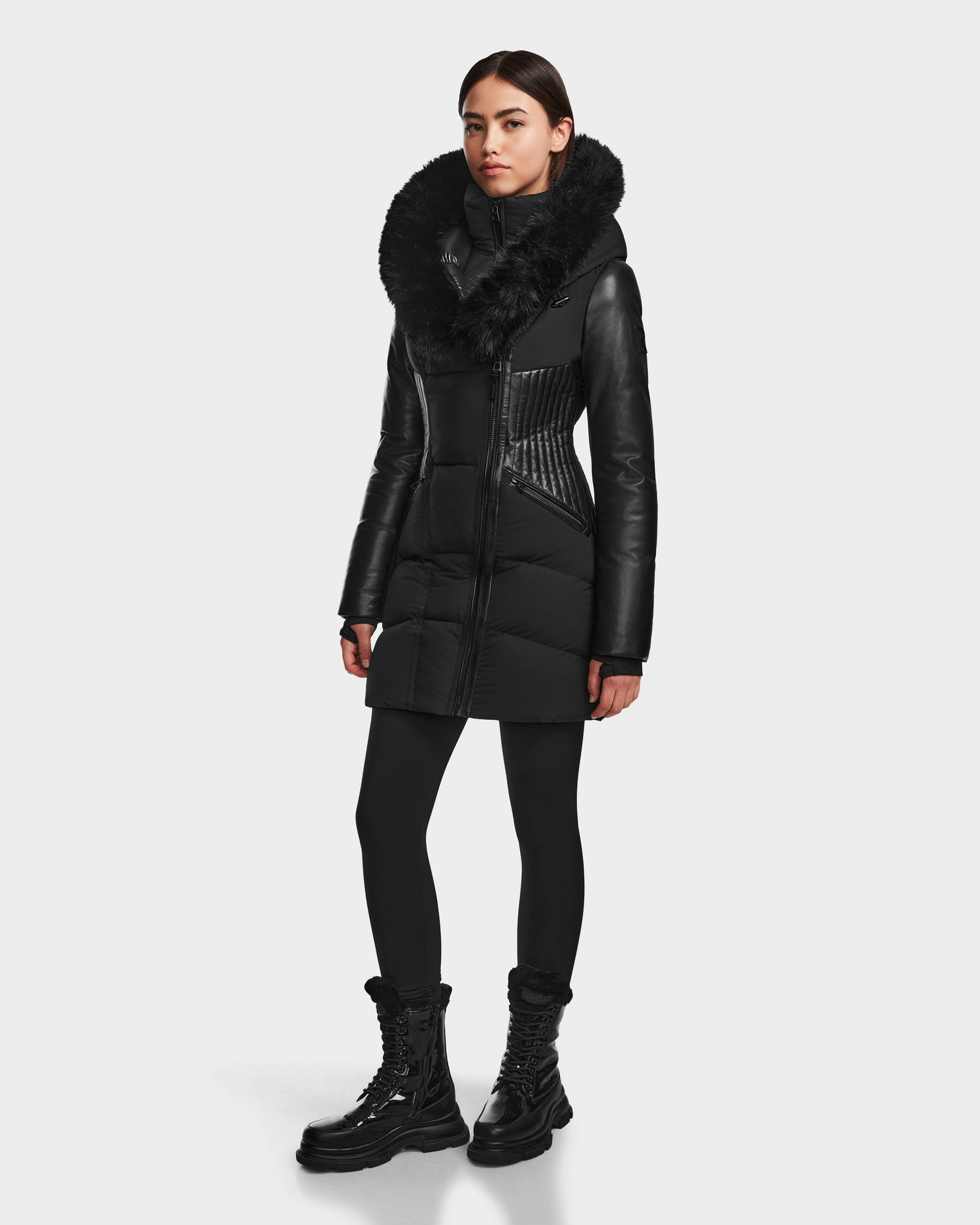 A woman models the RUDSAK SHAUNA - 8122586 BLACK winter coat with a large faux fur collar, paired with black leggings and lace-up boots against a plain white background.