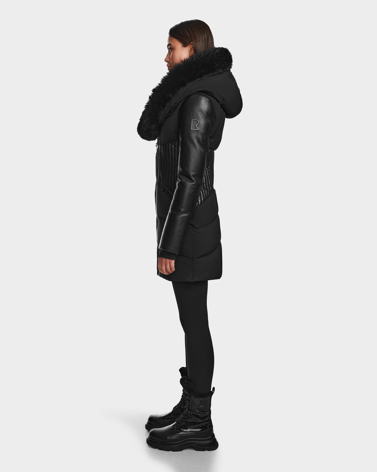 A woman in profile wears the RUDSAK SHAUNA - 8122586 BLACK quilted down puffer with a large fur collar, styled with leggings and black winter boots, posing against a plain white background.