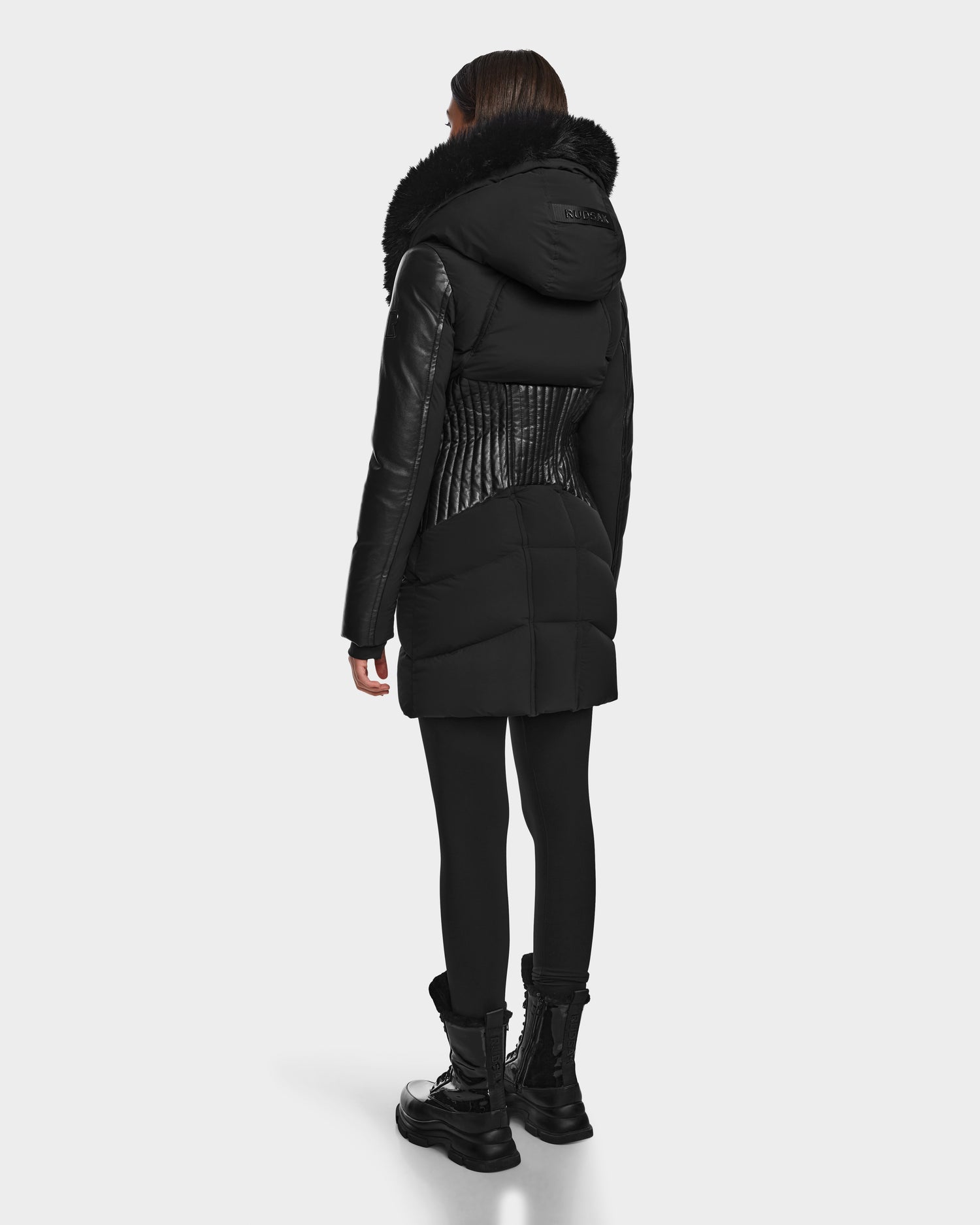 A person faces away, wearing the RUDSAK SHAUNA - 8122586 BLACK long quilted down puffer with a fur-lined hood, paired with black leggings and lace-up boots against a plain light background.