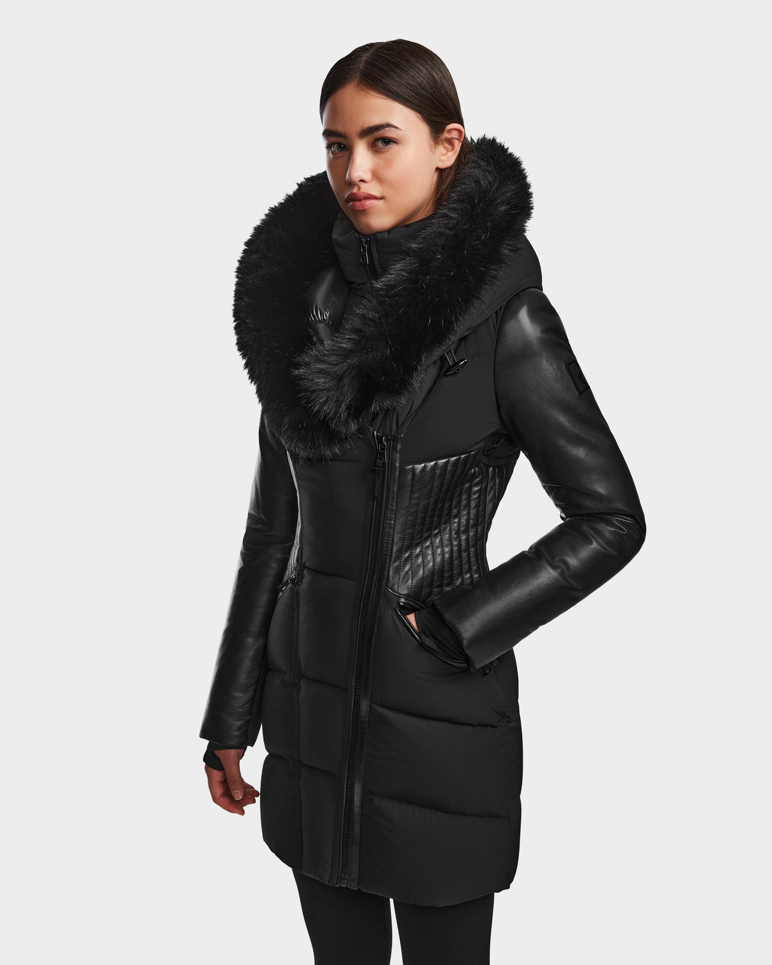 A woman models the RUDSAK SHAUNA - 8122586 BLACK quilted down puffer winter jacket with a large fur collar, standing against a plain white background with hands in pockets and looking slightly to the side.