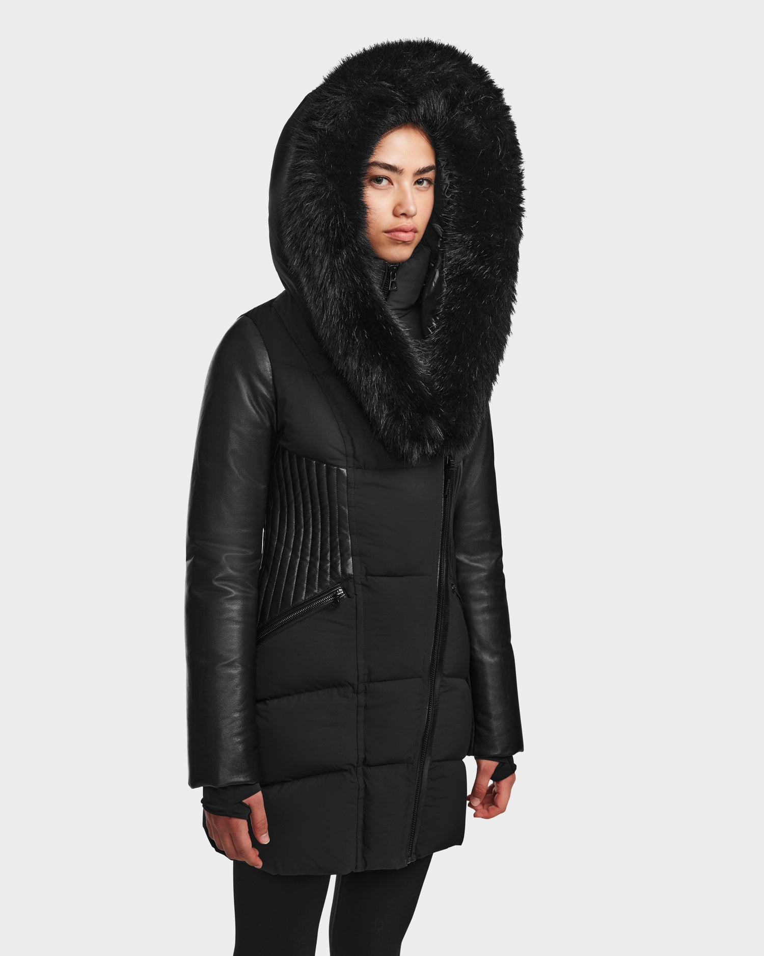 Wearing the RUDSAK SHAUNA - 8122586 BLACK puffer coat with quilted down, leather sleeves, and a faux fur-lined hood, a person stands against a plain light background.