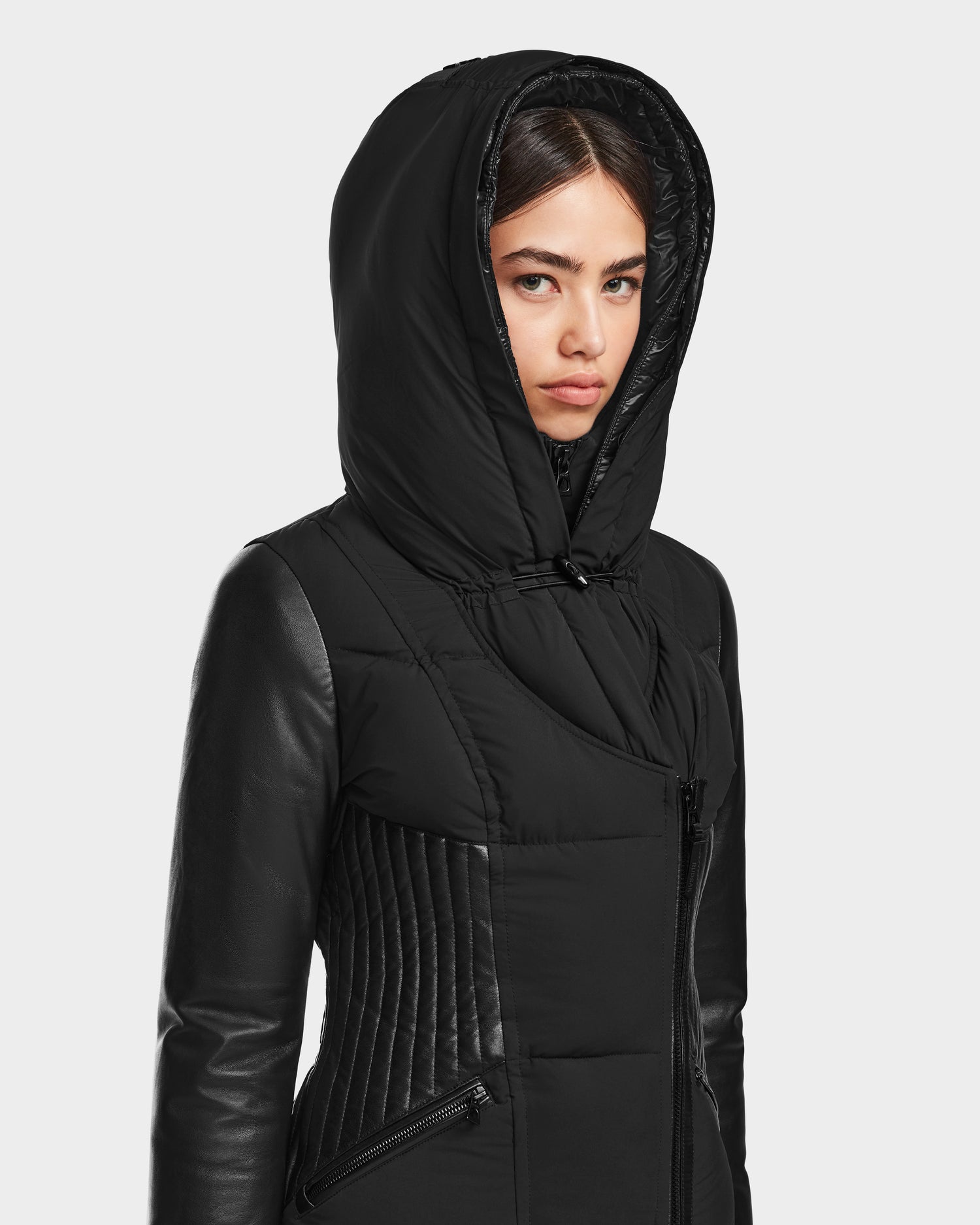 A young woman wears the RUDSAK SHAUNA - 8122586 BLACK women’s winter coat, featuring quilted and leather sections with RDS-certified down, looking ahead with a neutral expression against a plain light background.