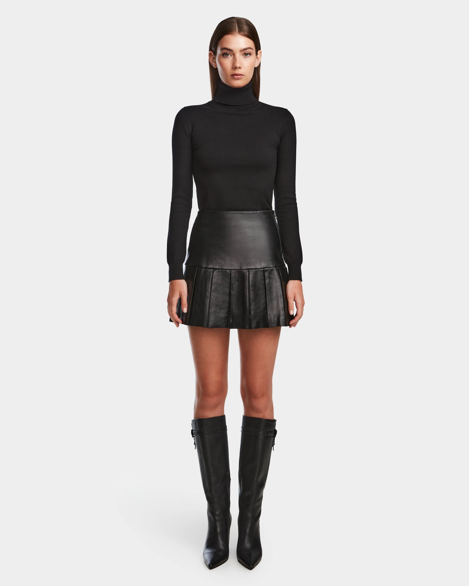 A woman faces forward against a white background, wearing a black turtleneck, knee-high black boots, and the Rudsak SOLANGE - 8524543 Black pleated leather skirt.