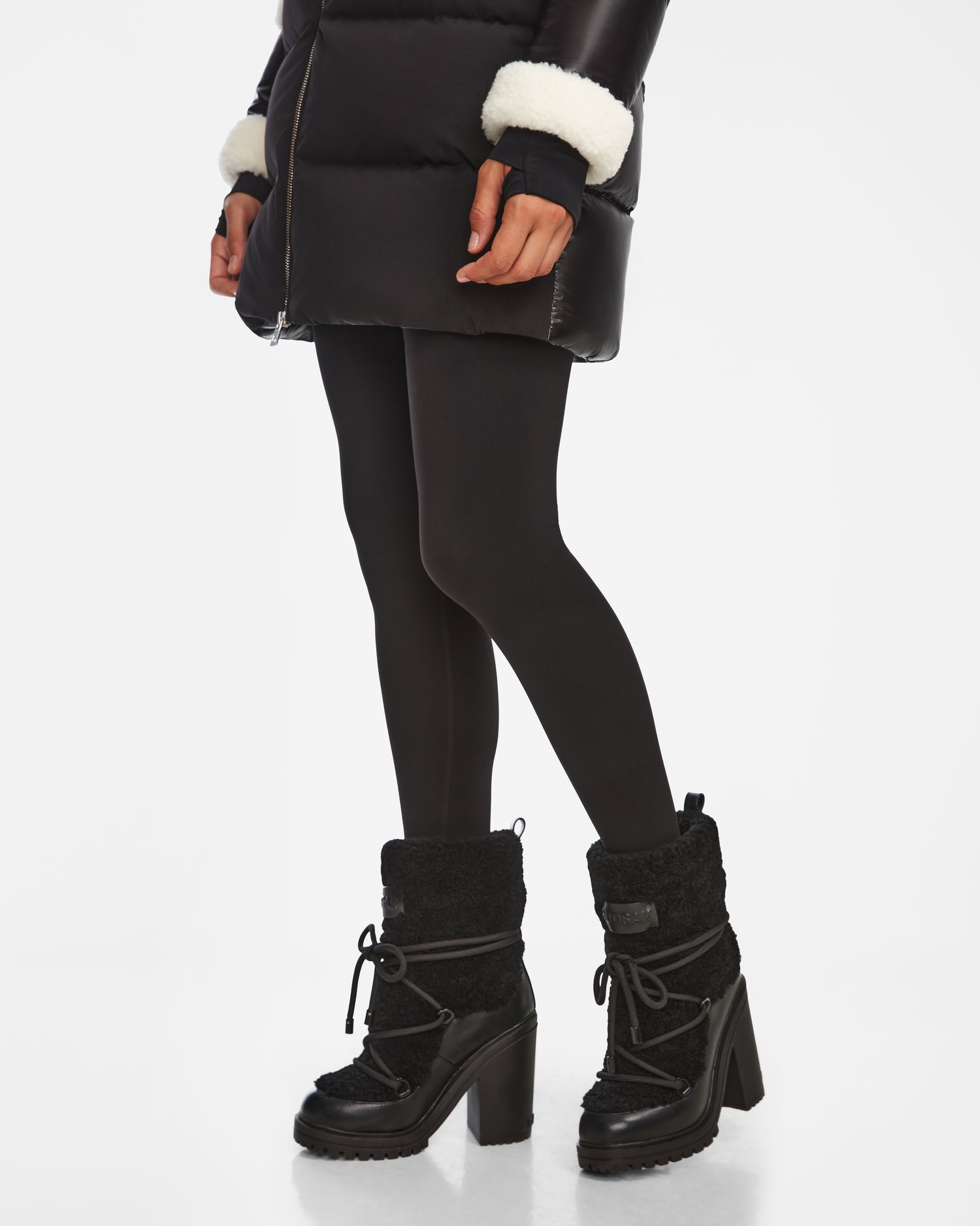 A person wears the Rudsak TALYNA - 8224504 Black puffer coat with white cuffs, black tights, and stylish women’s ankle boots, standing against a white background.