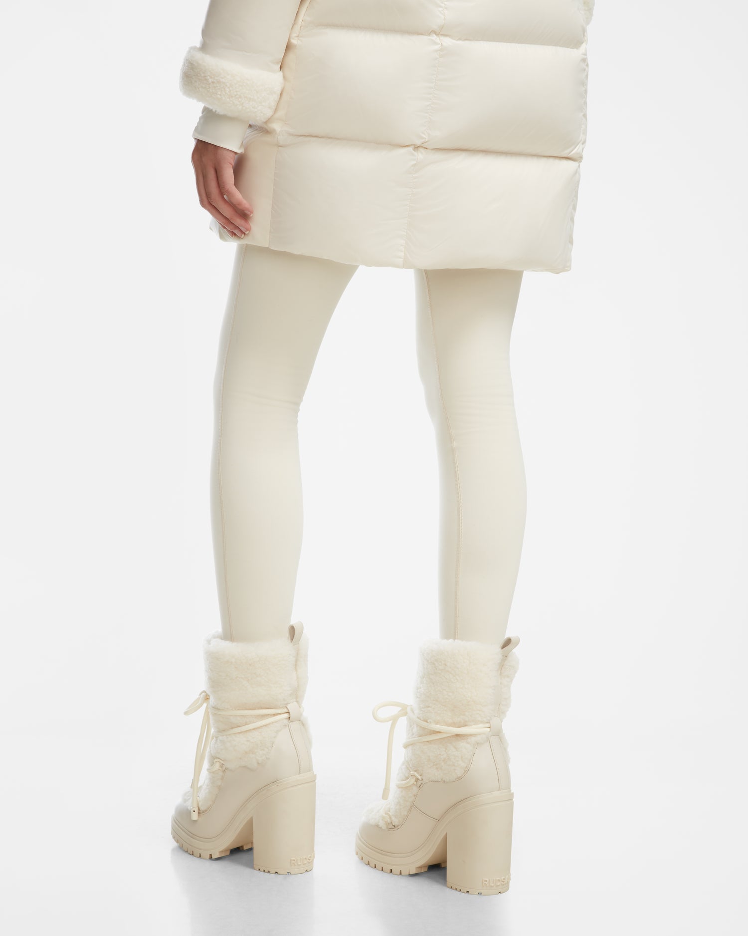 A person wears Rudsak TALYNA - 8224504 Iceland cream-colored leggings, a puffy cream coat, and womens high-heel block boots with faux fur trim, standing on a white background, shown from the back knee down.