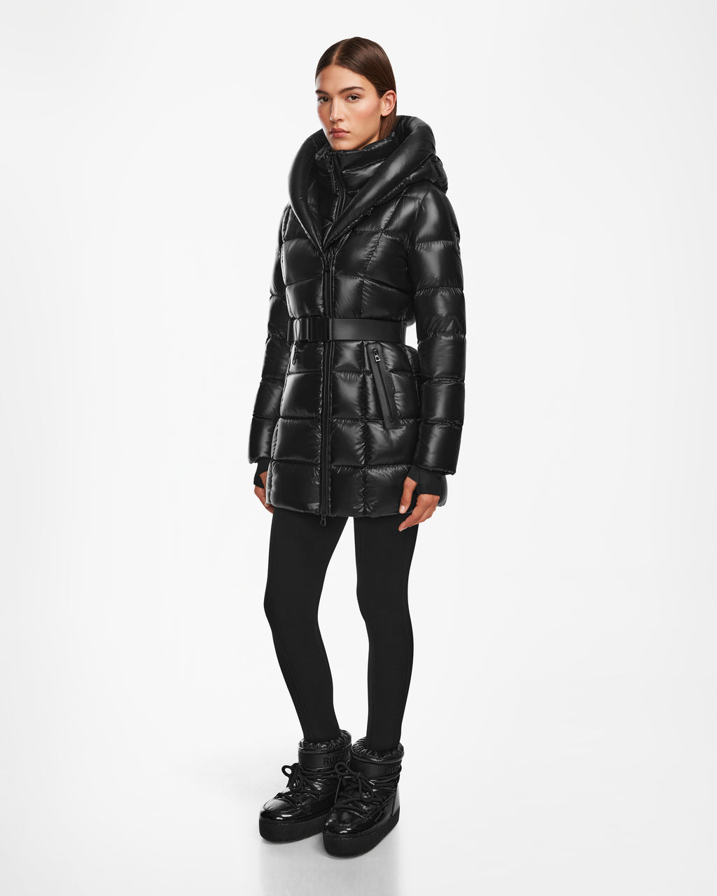 A woman stands against a white background wearing the Rudsak TAYLOR S - 8124820 Black, a shiny black womens down puffer jacket with a high collar, hood, belted waist, and quilted stitching. She pairs it with black leggings and boots.