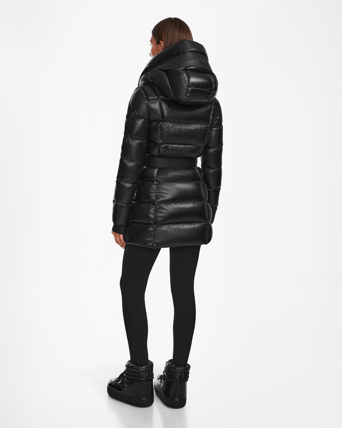 A woman faces away, wearing Rudsak’s TAYLOR S - 8124820 Black shiny water-repellent hooded winter jacket, with black leggings and platform shoes, set against a plain white background.