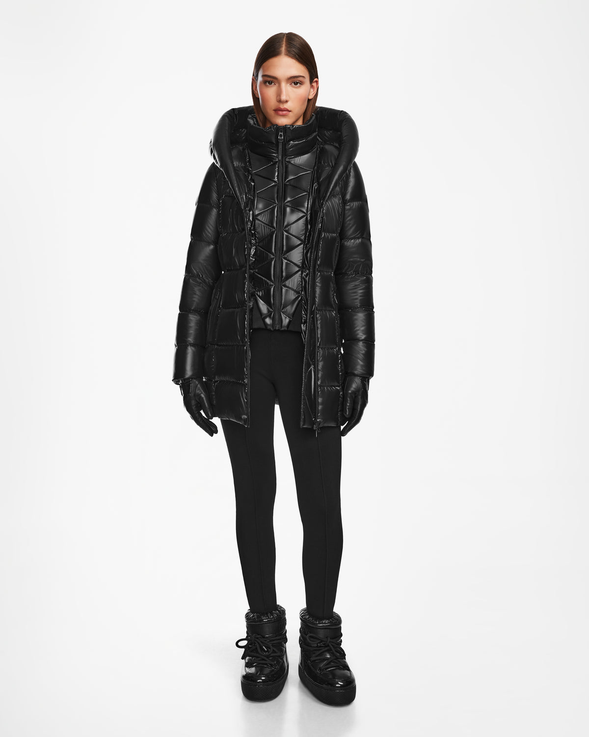 A woman with straight brown hair and a neutral expression models the Rudsak TAYLOR S - 8124820 Black water-repellent womens down puffer jacket with a hood, paired with black leggings, gloves, and winter boots against a white background.
