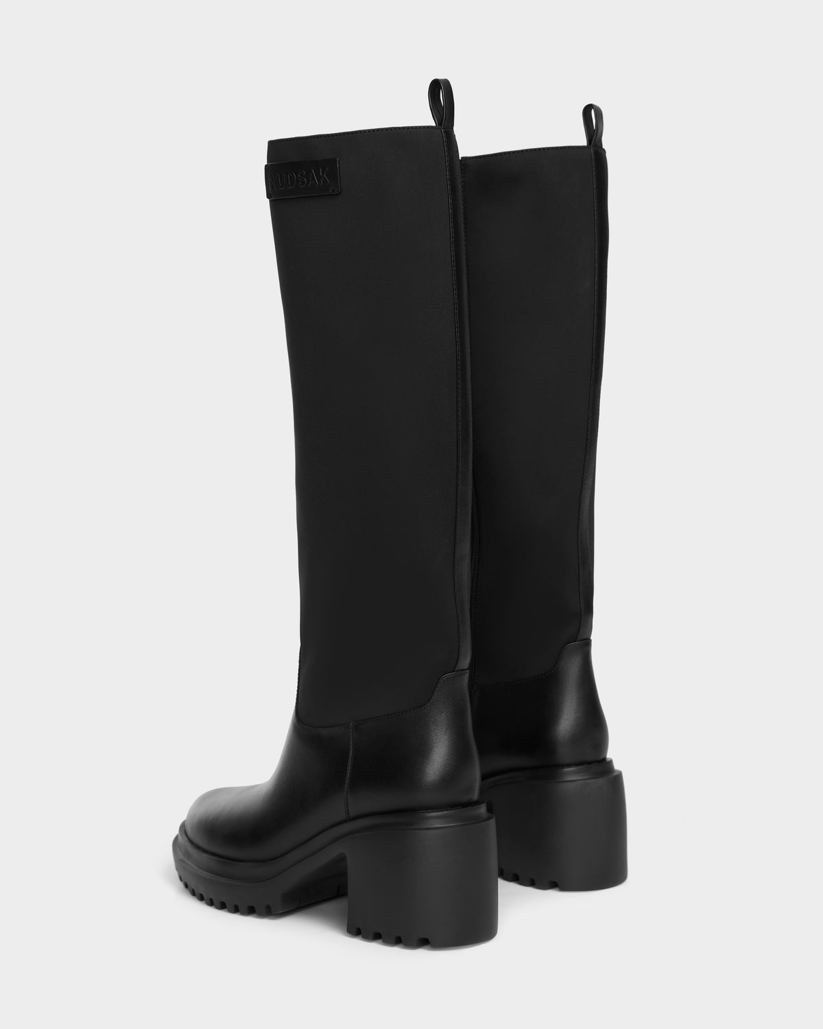 The Rudsak TIARA - 8224529 Black boots feature a high shaft, chunky heels, and textured soles, displayed from a rear side angle against a plain light background.