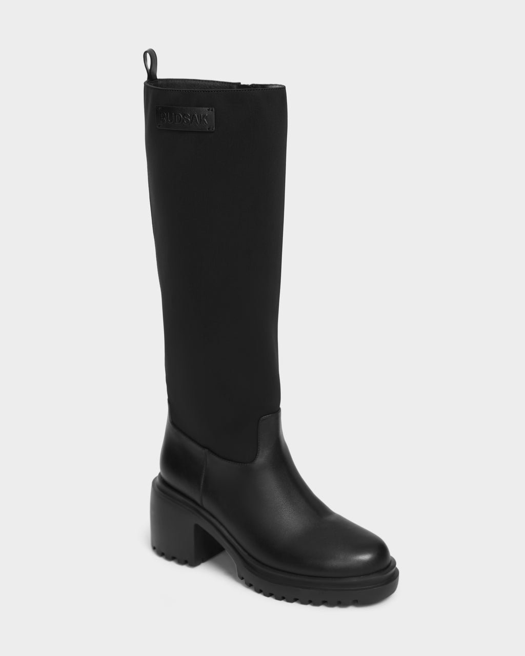 The Rudsak TIARA - 8224529 Black is a womens mid-heel, high-shaft boot with a chunky heel, lug sole, pull tab, and subtle brand patch near the top. Shown against a plain white background.