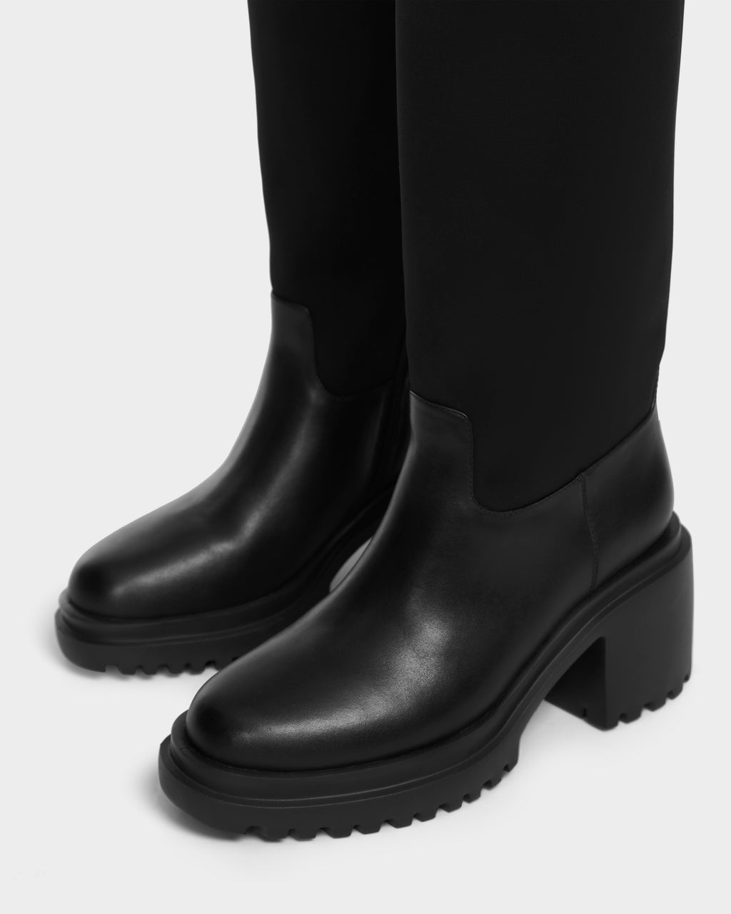 A close-up of the Rudsak TIARA - 8224529 Black boots features chunky, textured soles and a high block shaft, shown on a person wearing black pants.