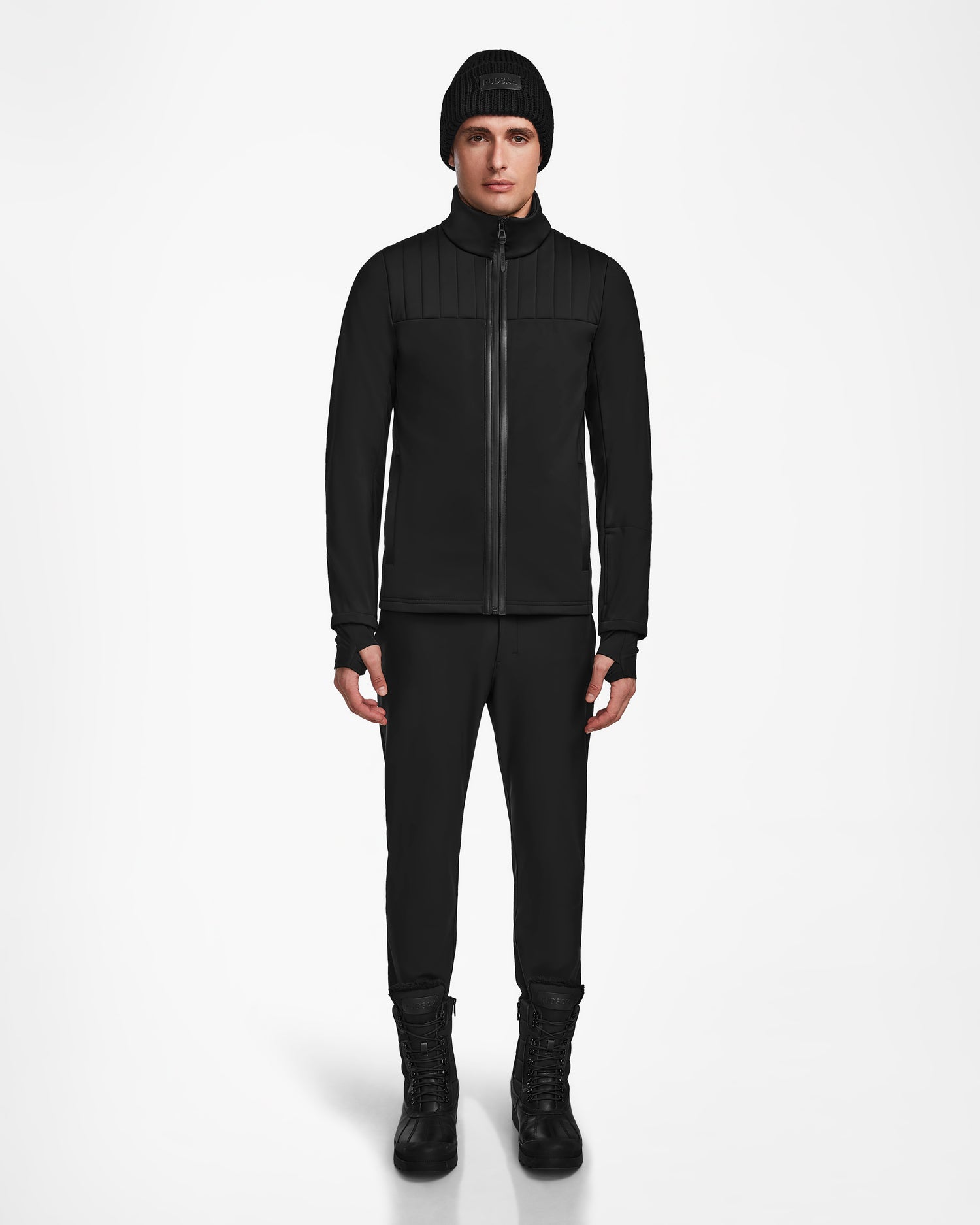 A person stands facing forward, wearing the Rudsak TREVOR - 6124519 Black mens ski jacket, black pants, and black lace-up boots against a plain white background.