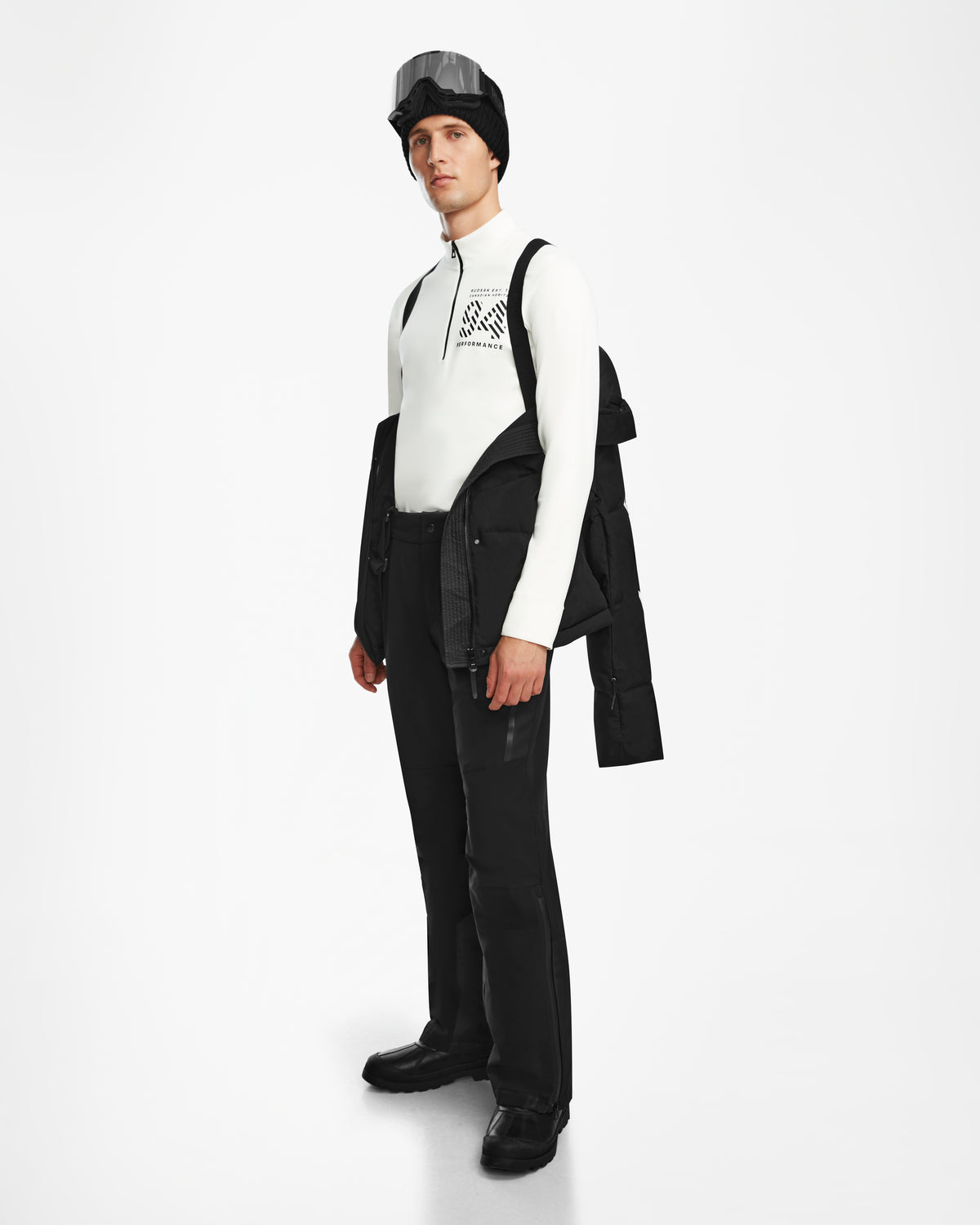 A man stands against a white background wearing the Rudsak USHER - 6124516 Black helmet, a water-resistant ski jacket over his shoulders, a white geometric-pattern zip-up top, black pants, and black shoes.